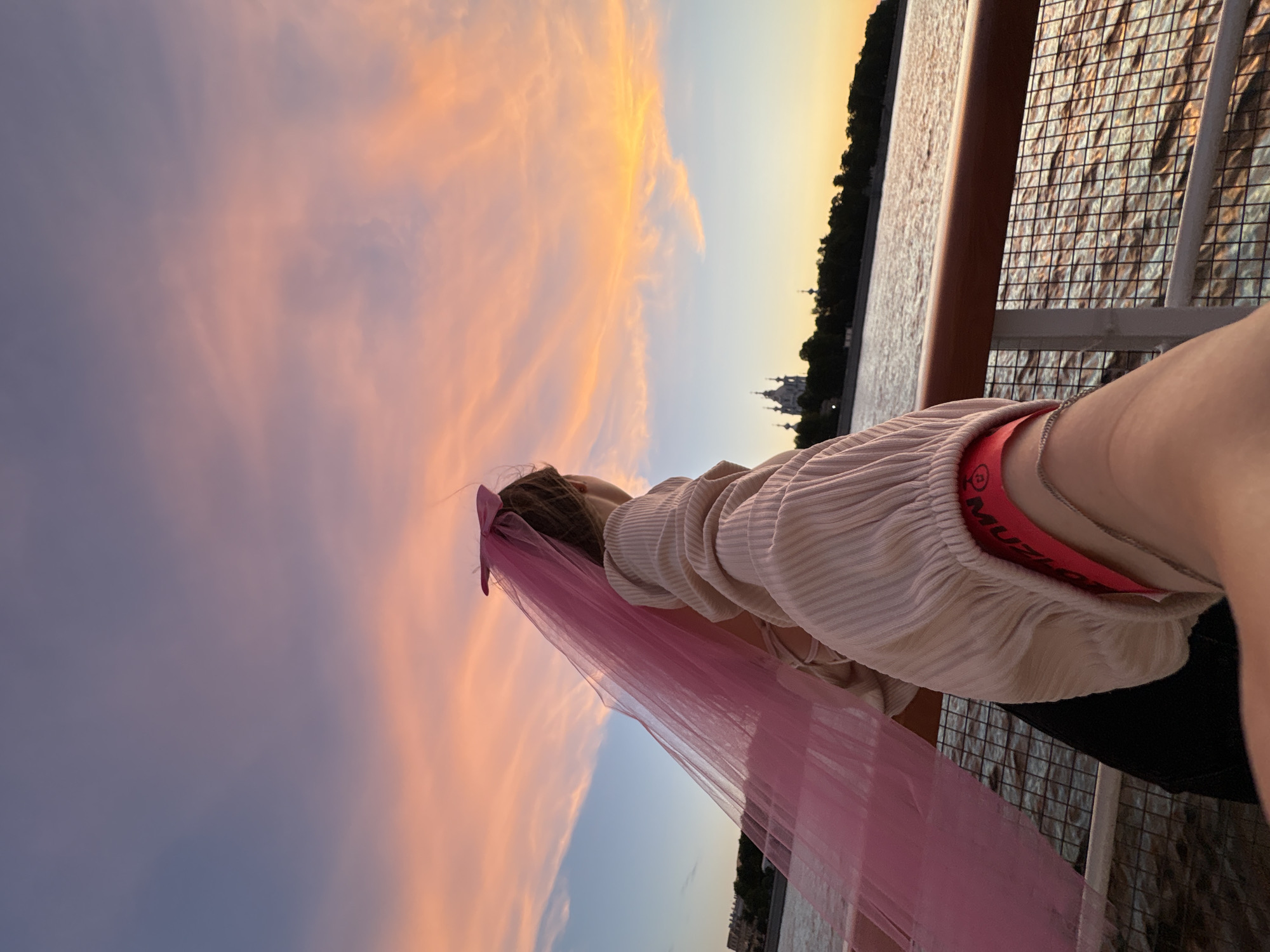A person wearing a pink veil and a ribbed beige shirt stands on a boat at sunset. The person is looking out at a body of water, with a distant cityscape visible on the opposite bank. The sky is filled with vibrant orange and pink clouds. The railing of the boat is in the foreground, with the water reflecting the sunset. The cityscape includes several domed buildings, characteristic of Saint Petersburg.
