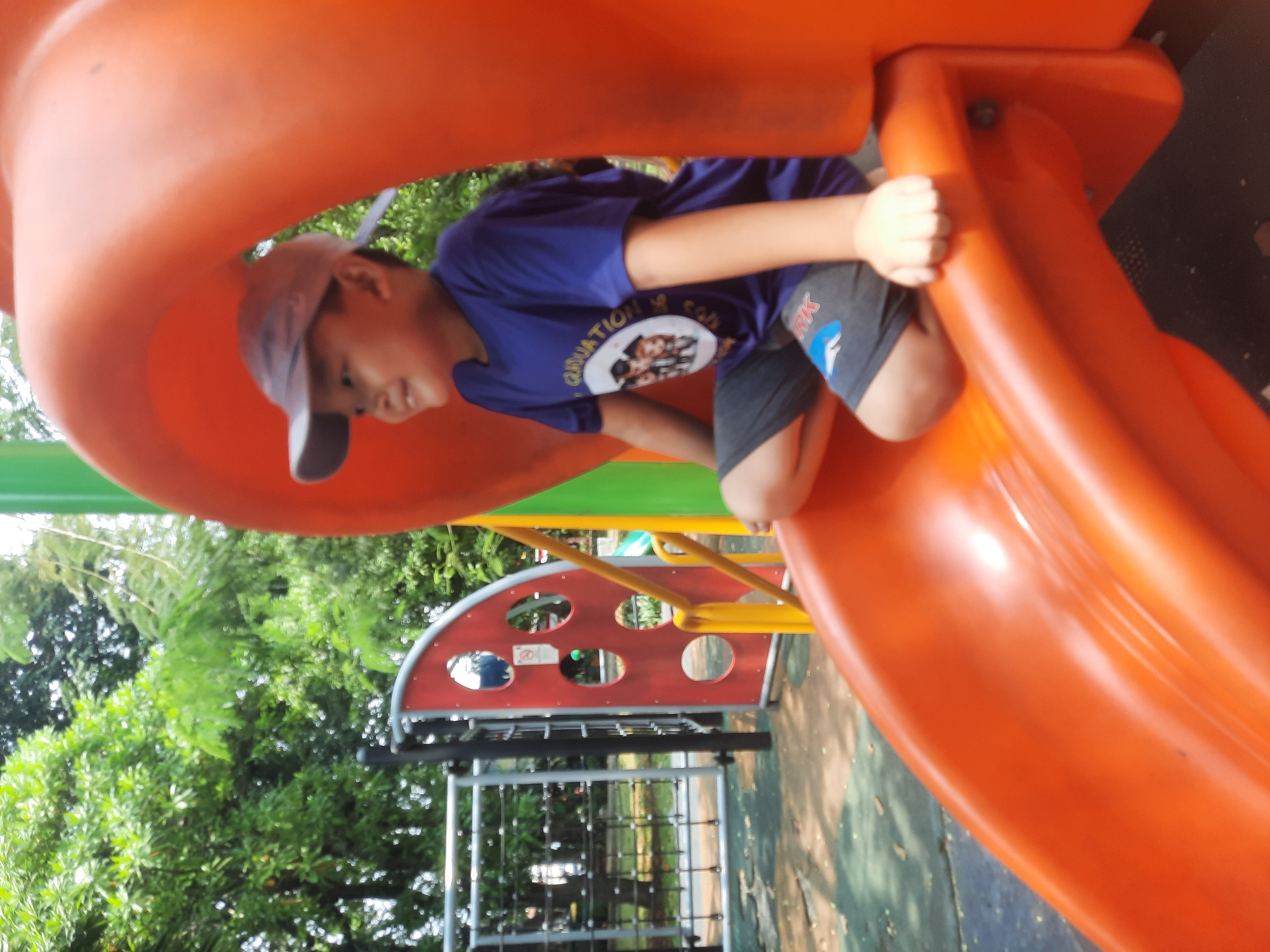A young Asian boy wearing a gray baseball cap, a purple t-shirt with a graduation emblem, and gray shorts sits on an orange plastic playground slide. The slide is part of a larger playground structure visible in the background, which includes red panels with circular cutouts, yellow railings, and green and blue elements. The boy is smiling and looking to his right, with his hands resting on the slide. The ground beneath the slide appears to be a dark, possibly rubberized, surface. Lush green trees and foliage form the backdrop, suggesting an outdoor setting in a park or playground. The lighting suggests it is daytime. The context of North Jakarta, Indonesia, implies this is a typical playground found in an urban park within that region.