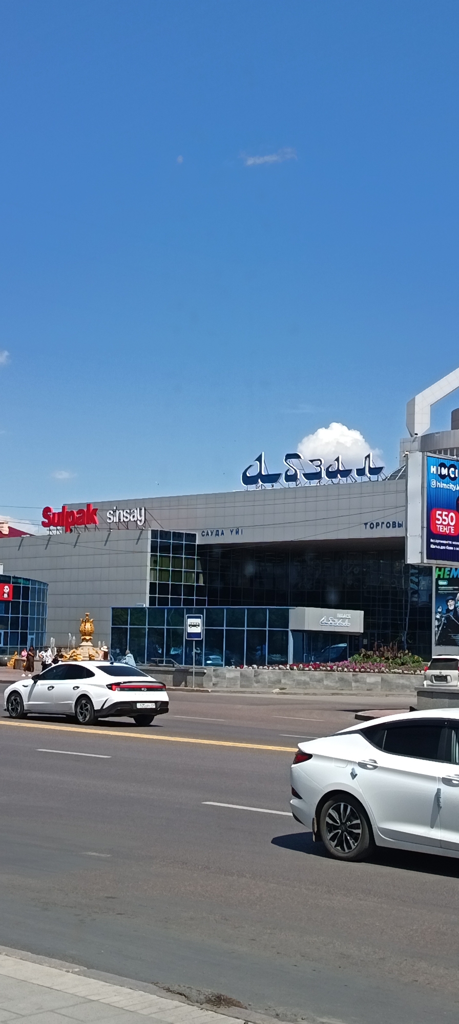 A wide, vertical shot shows the facade of a modern shopping center in Qarağandı, Kazakhstan. The building is light-colored with large, dark-tinted windows. Above the windows, several prominent signs are visible. On the left, a red sign reads "Sulpak" in a stylized font, followed by "sinsay" in white text. To the right of this, a large blue Arabic script sign is present. Further to the right, more signs are visible: "САУДА ҮЙІ" (Sauda Üi, meaning Shopping House in Kazakh), "ТОРГОВЫ" (Torgovy, meaning Commercial in Russian), and a blue and white billboard with text including "HIMCI" and "550 ТЕНГЕ" (550 Tenge, the currency of Kazakhstan).
In the foreground, a paved road with yellow and white lane markings is visible. Two white sedans are on the road. The one on the left is a modern sedan, parked or moving slowly in the right lane. The one on the right is closer to the camera and is in the left lane, also a modern sedan.
On the sidewalk in front of the shopping center, there are several people walking and standing. A golden fountain structure with figures is located to the left of the main entrance. Planters with greenery and flowers are also visible along the base of the building. The sky is a bright blue with a few small, scattered white clouds and a faint white object, possibly the moon. The overall impression is a bright, sunny day in an urban setting.
