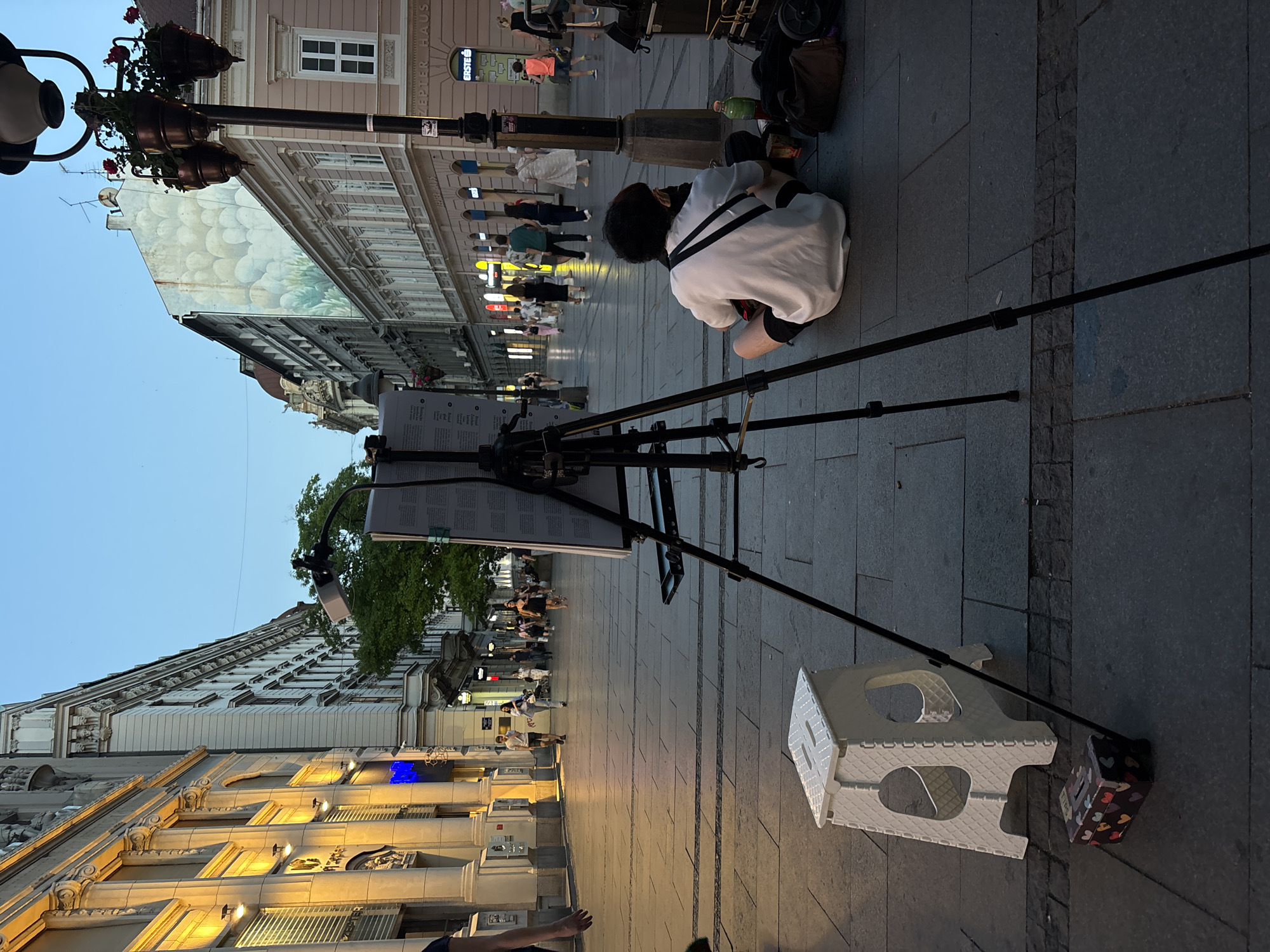 The image depicts a street scene in Belgrade, Serbia, possibly in the evening or early morning given the lighting. A young person wearing a white hoodie and black shorts is sitting on the pavement, facing away from the camera. They appear to be engaged with a portable easel and a tablet or display board mounted on it. A small white folding stool is placed nearby, along with a decorative box with heart patterns.
In the background, people are walking along the pedestrian street, some in groups and others individually. The street is lined with buildings showcasing architectural details, with lights from shopfronts and streetlights illuminating the scene. The sky is a pale blue, suggesting either dawn or dusk.
The overall atmosphere seems to be one of quiet activity on a city street. The subject's posture and focus suggest concentration on their work or study. The presence of other pedestrians indicates a public space that is active even at this time. There is some visible text on a sign attached to the easel, but it is not legible. The buildings have ornate facades, some with illuminated windows.