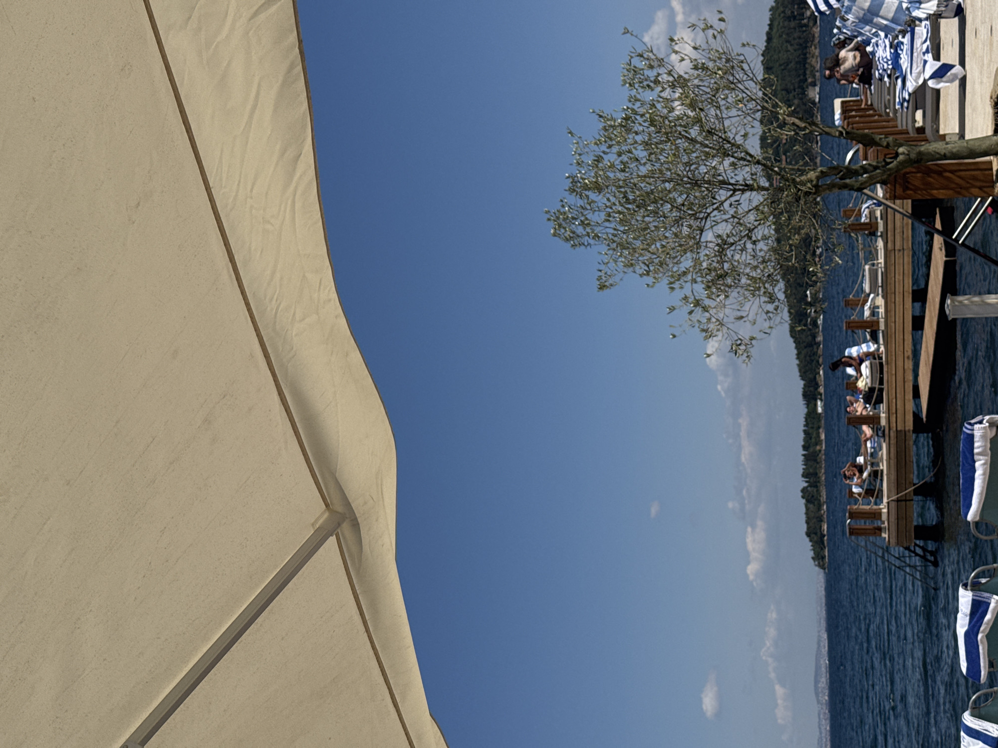 A wide-angle, low-angle shot captures a scene at a waterfront resort on one of the Prince Islands in Turkey. In the foreground, a large, cream-colored umbrella partially obstructs the view of the bright blue sky. To the right, a small, leafy tree stands tall, its branches reaching towards the sky.
In the mid-ground, a wooden pier extends into the deep blue water. Several people are seen relaxing on lounge chairs on the pier. Some are lying down, while others are sitting up. Blue and white striped towels are draped over some of the lounge chairs. A set of metal stairs leads down from the pier into the water.
In the background, a distant shoreline is visible, characterized by a densely wooded island with rolling hills. The water is calm, with gentle ripples on the surface. The overall impression is one of a peaceful and relaxing day at a seaside resort.