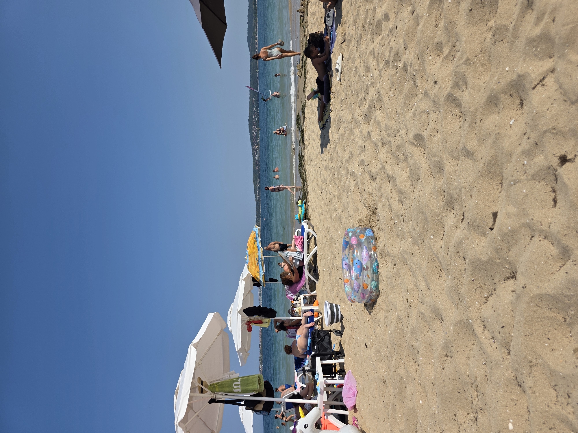 The image shows a sunny day at a beach in Chernomorets, Bulgaria. The sand is light brown and scattered with small shells and debris. Several beach umbrellas provide shade for beachgoers. In the foreground, an inflatable kiddie pool with fish designs is placed on the sand.
Various individuals are visible enjoying the beach. In the middle ground, people are lounging on chairs and sunbeds, some reading or resting. Others are in the shallow water, swimming or wading. A woman in a light-colored swimsuit walks out of the water towards the shore, carrying a hat. Further out in the sea, a person is windsurfing. In the background, a hilly coastline dotted with buildings is visible under a clear blue sky. The sea is calm and a light blue color.