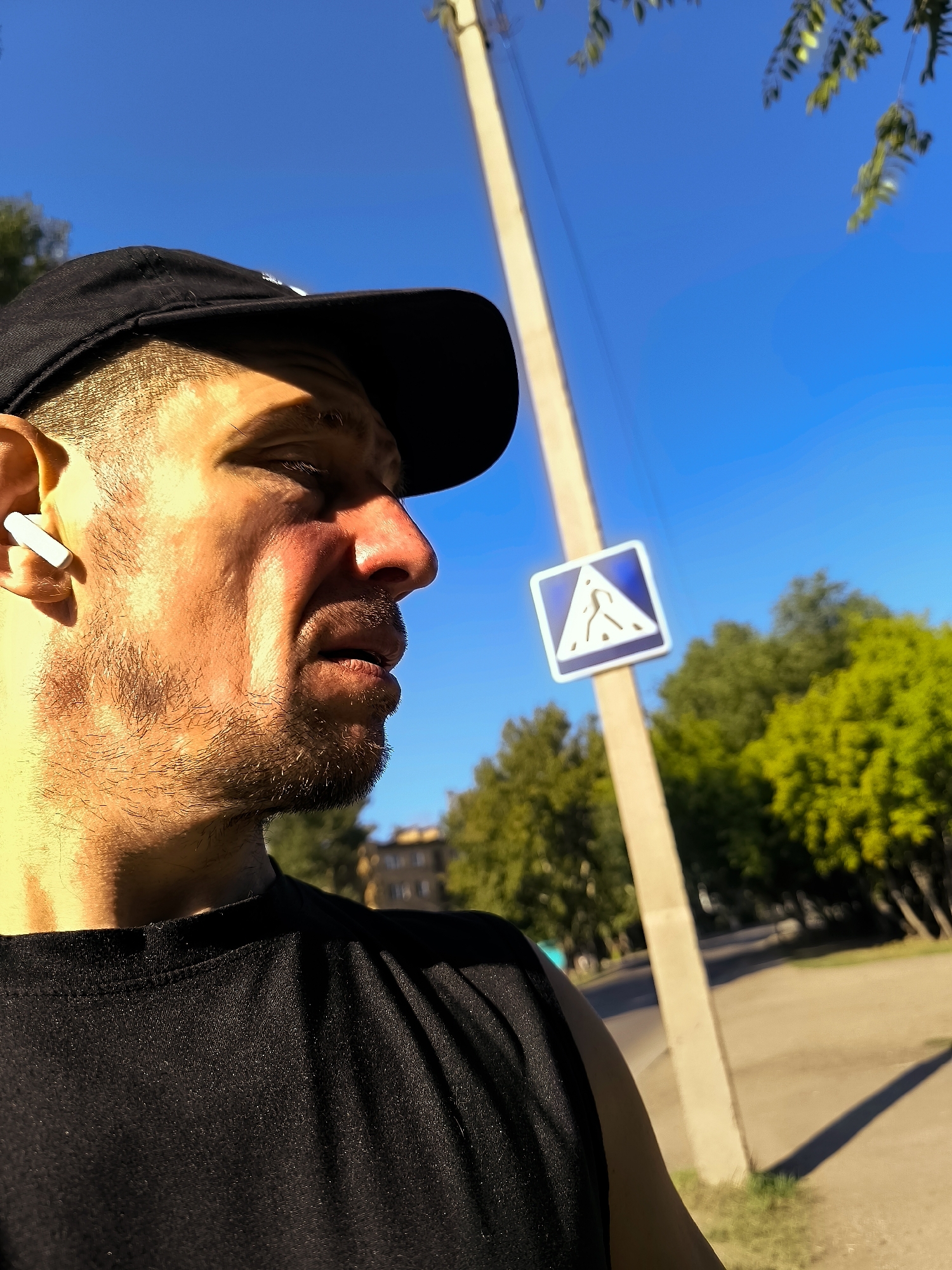 A medium shot captures a man from the side, looking to the right, with a black baseball cap on and a white earbud in his left ear. He is wearing a black t-shirt. Behind him, a light-colored pole with a blue and white pedestrian crossing sign is visible. The background shows green trees and a clear blue sky, suggesting an outdoor setting during the day. The image appears to be taken in the city of Qarağandı, Kazakhstan, as implied by the administrative context of the request.