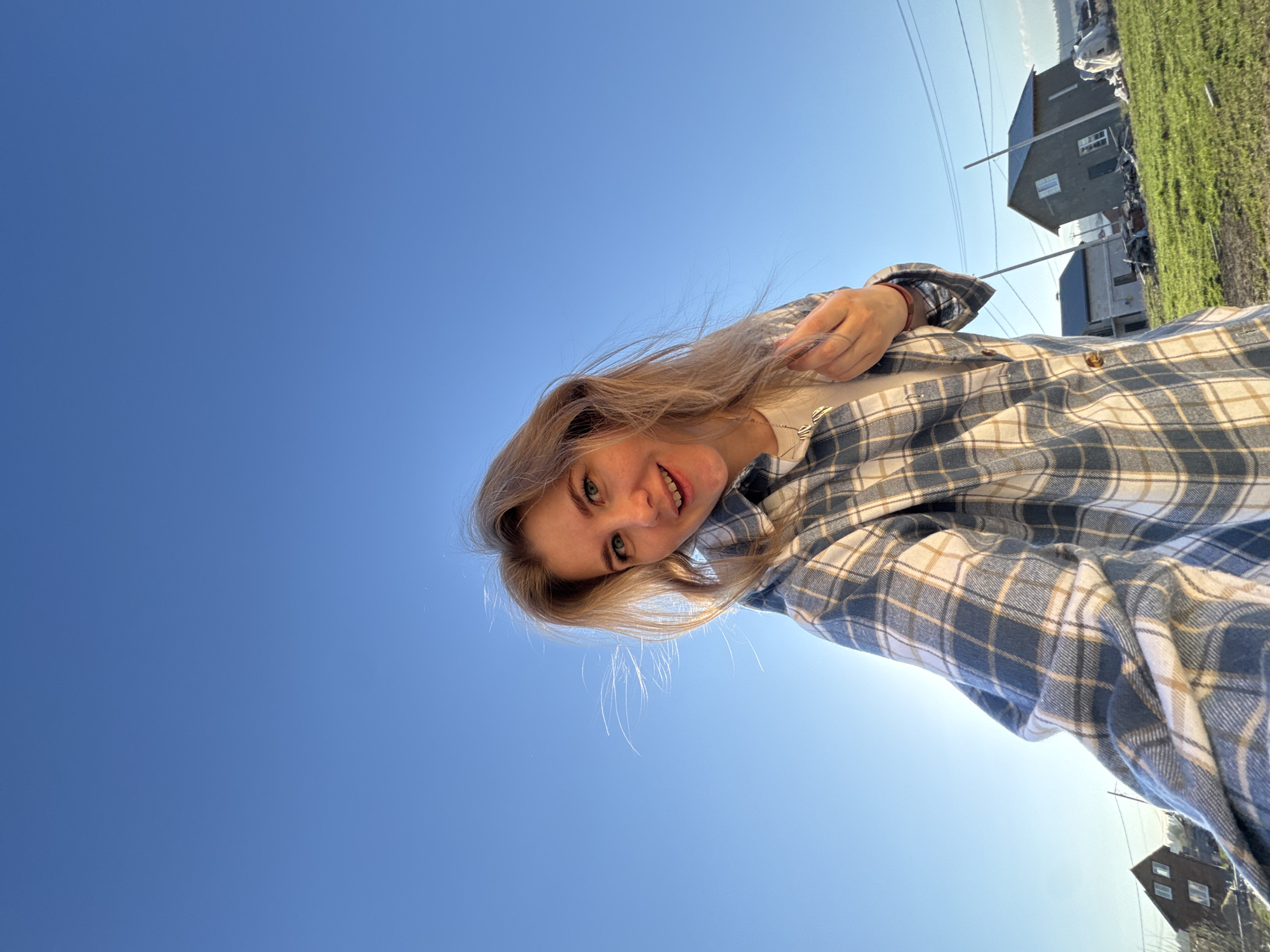 A woman is pictured from a low angle, looking up towards the camera. She has blonde hair and is wearing a plaid shirt. Behind her is a clear blue sky and a landscape with buildings and green fields, which are located in Ферма, Russia. Power lines are visible overhead.