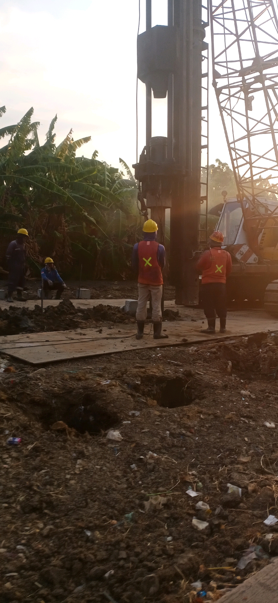 The image depicts a construction site in Surabaya, Indonesia. In the foreground, disturbed earth with scattered debris and several shallow holes is visible. A wooden platform lies on the ground, extending towards the center of the frame. Further back, three construction workers are present. One worker, wearing a blue jumpsuit and a yellow hard hat, stands to the left, facing away from the camera. Another worker, wearing a blue jacket and a yellow hard hat, is crouched down near the center, also facing away. A third worker, in the center of the frame and facing away, wears a red vest with a yellow "X" symbol and a yellow hard hat. To the right of this worker, another construction worker, also facing away, wears an orange vest with a yellow "X" symbol and a yellow hard hat. Behind the workers, a large piece of heavy machinery, possibly a pile driver, dominates the background. The sky above is bright, suggesting it is either sunrise or sunset, with sunlight casting long shadows. Lush green banana trees are visible on the left side of the image, indicating the presence of vegetation. The overall scene is consistent with ongoing construction work.