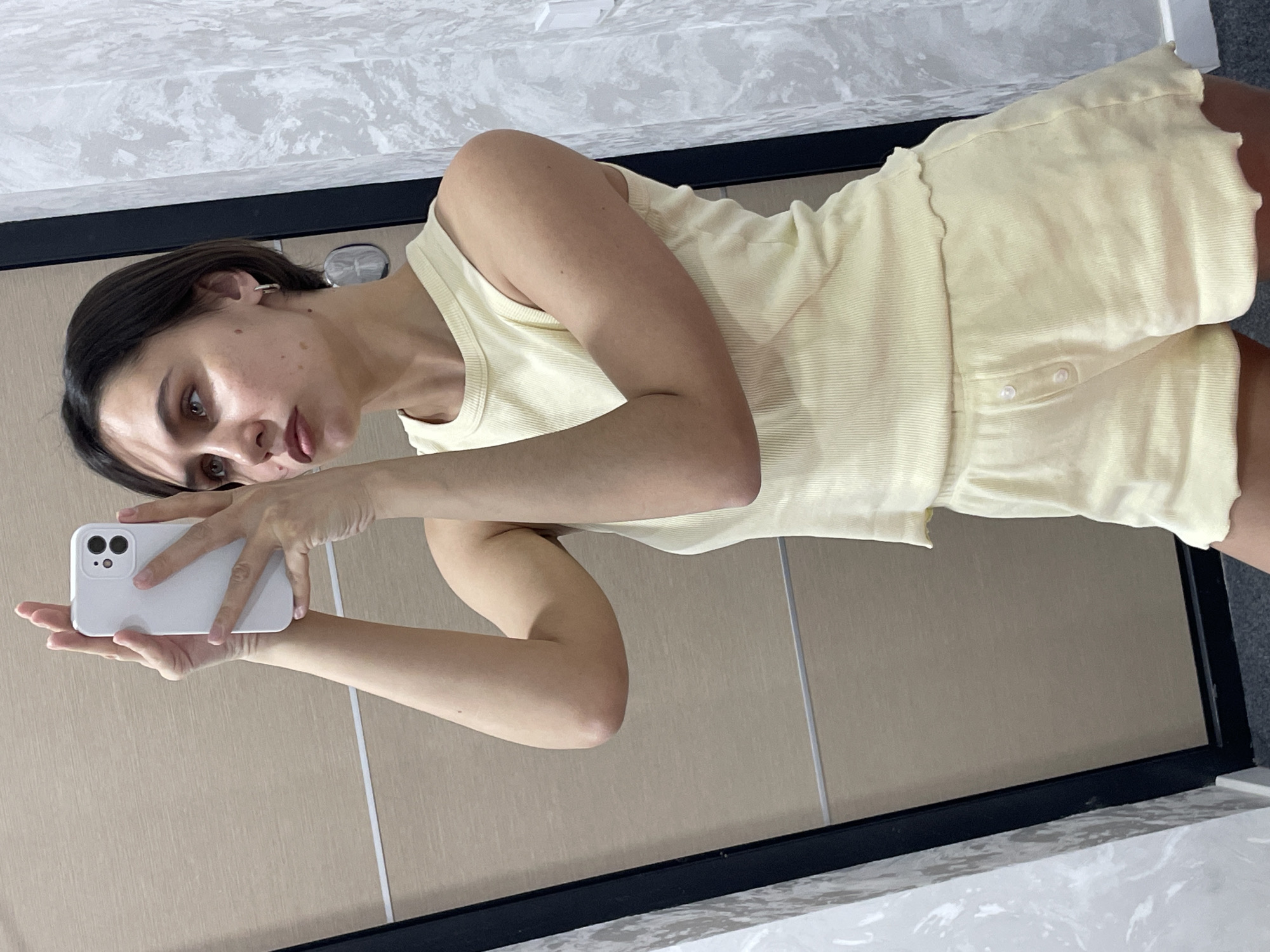 A woman is taking a selfie in front of a mirror. She is wearing a light yellow ribbed tank top and matching shorts with buttons. She is holding a white smartphone. The background shows a wall with beige tiles and a countertop with a gray and white marble pattern. The location is likely inside a home or apartment, but its location in Izhevsk, Russia, cannot be determined from the image.