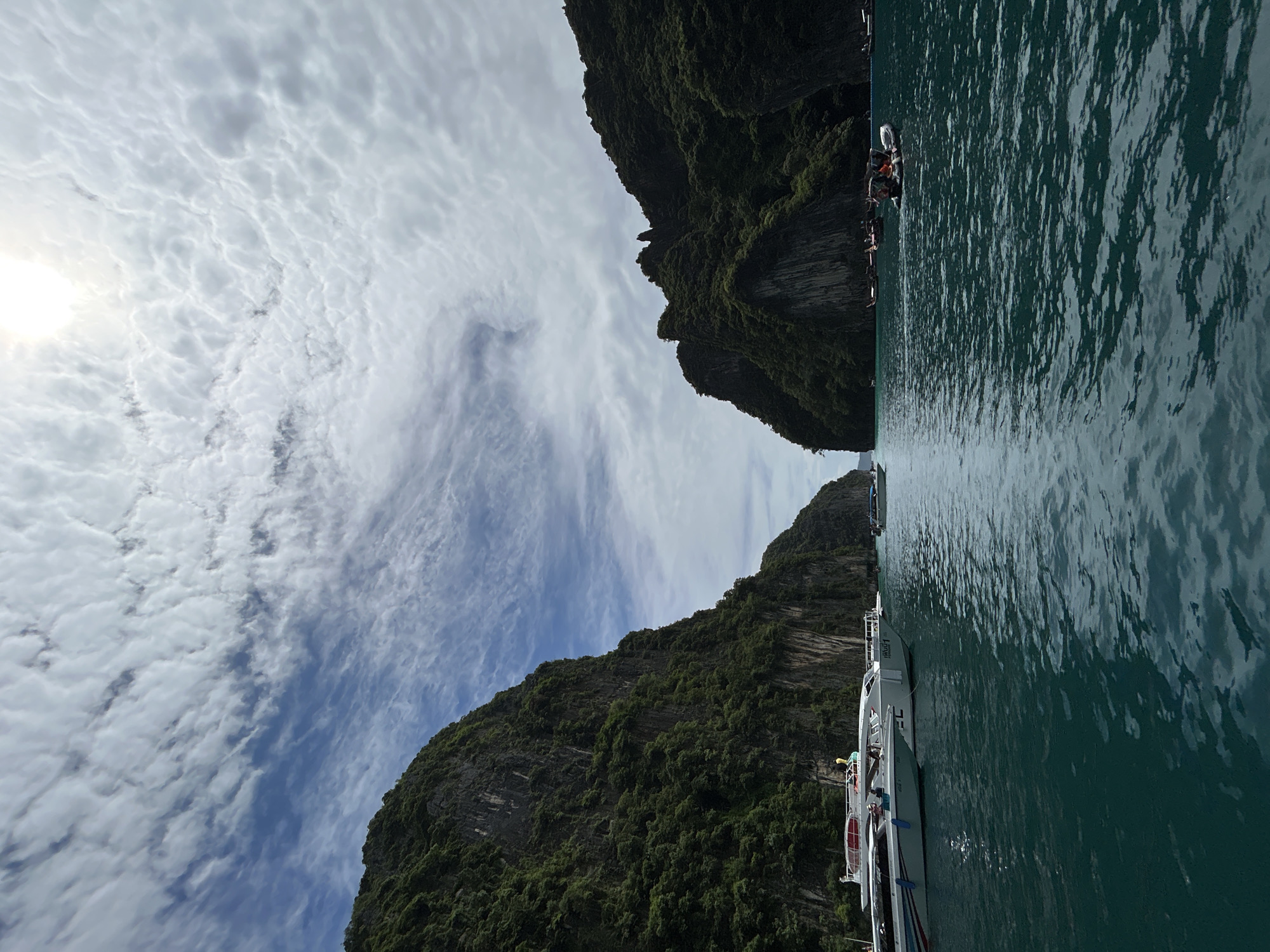 In a picturesque coastal scene in Unknown, Thailand, a body of water, likely the Andaman Sea, reflects the bright sky with scattered, fluffy white clouds and a visible sun in the upper right quadrant. Towering, lush green limestone cliffs flank the waterway, creating a dramatic and natural passage. Several boats are present: a line of white speedboats, some with passengers, are docked on the left, while smaller longtail boats are visible further in the distance on the right. A small, inflatable boat with people aboard navigates the water closer to the viewer. The overall impression is one of a serene and popular tourist destination, showcasing the natural beauty of the Thai coast.