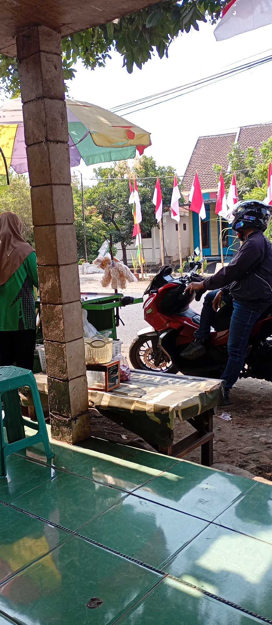 The image depicts a street-side stall in Pendil, Indonesia, possibly during a local celebration given the numerous Indonesian flags displayed. The stall features a brick pillar in the foreground, supporting an awning. To the left, a person wearing a brown hijab and a green shirt stands near some products. A red motorcycle is parked to the right, with a person wearing a black helmet and jeans seated on it, appearing to be a customer or passerby. Various items are displayed on tables, including snacks and what appear to be baskets. In the background, houses with tiled roofs and trees are visible, along with more flags. The ground is tiled green in the foreground, and a dirt path is visible further back. The lighting suggests it is daytime.