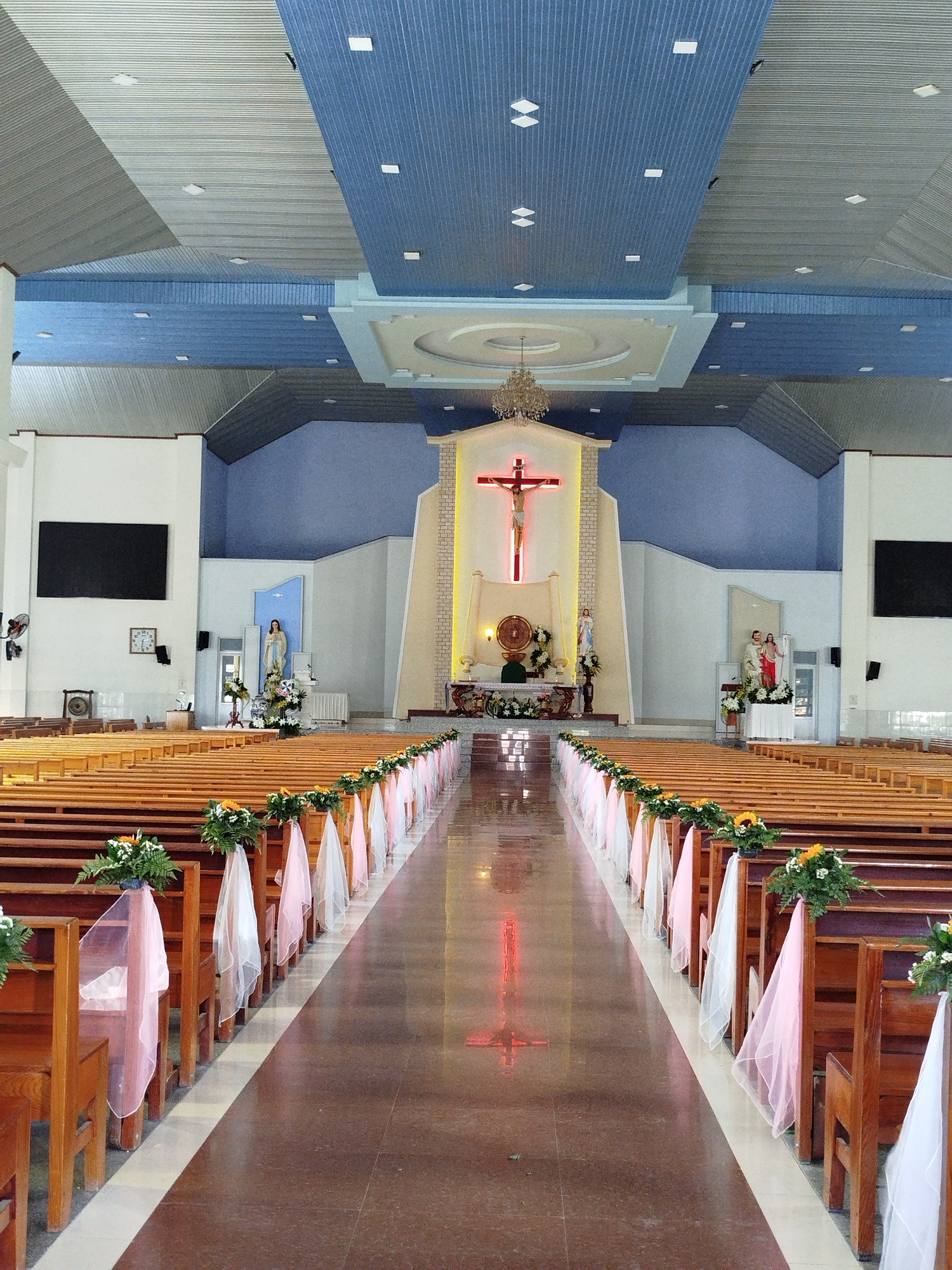The image shows the interior of a church in Xuân Thượng, Vietnam. Rows of wooden pews are decorated with floral arrangements and pink and white ribbons. The aisle leads to the altar, which features a cross illuminated with red light. Statues of religious figures are visible on either side of the altar. The walls are painted white and blue, and the ceiling is a combination of flat and vaulted sections with recessed lighting. A chandelier hangs above the altar. Black screens are mounted on the walls on either side of the sanctuary. A clock is visible on the left wall. The floor is polished and reflects the red light of the cross.