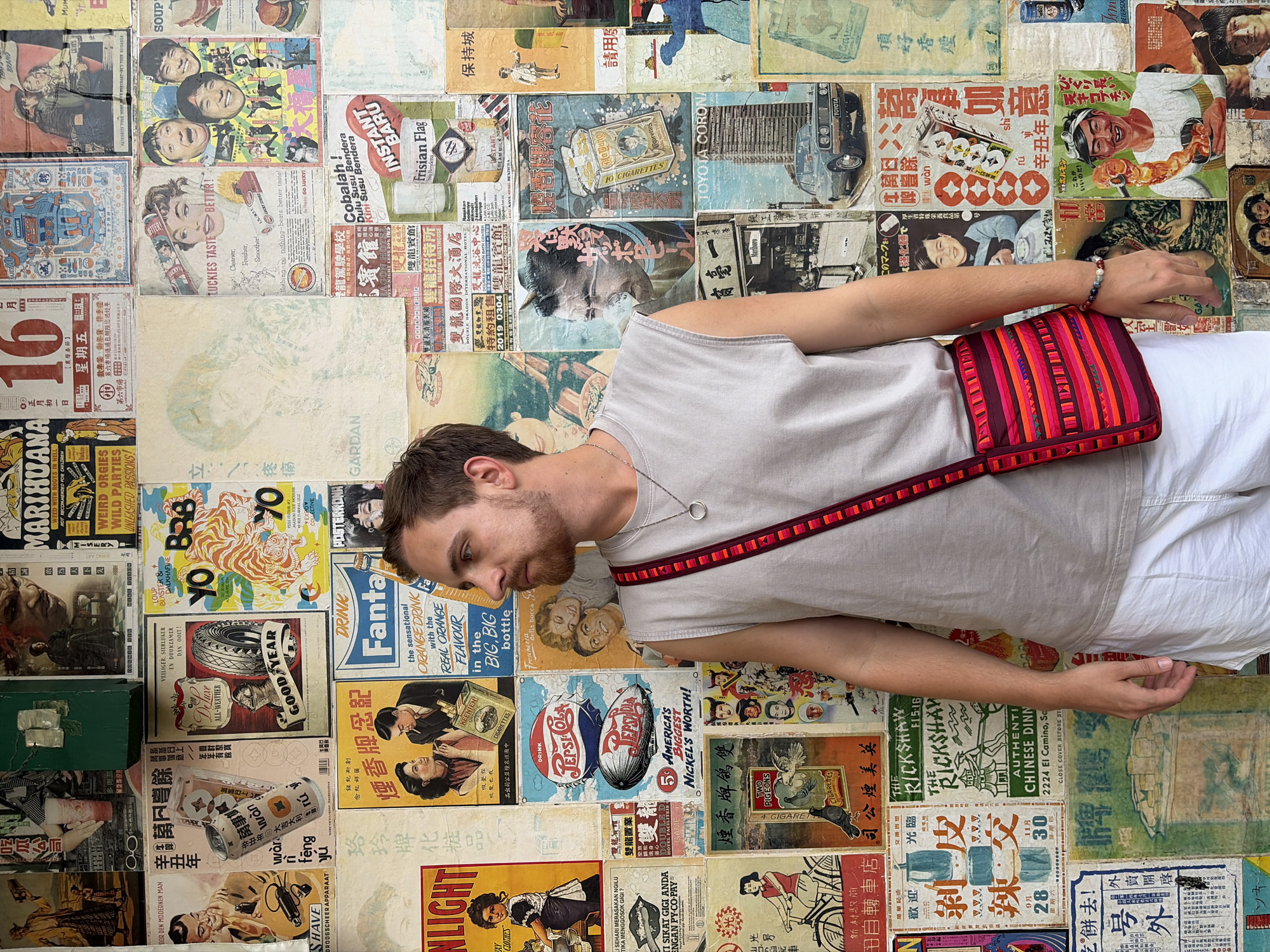 A person wearing a light gray tank top and white shorts stands in front of a wall covered in vintage advertisements and posters. The person has a dark shoulder bag with red and orange striped patterns. The posters feature a mix of Asian and Western advertisements, including brands like Fanta, Pepsi-Cola, Toyota, Marlboro, and several local brands in Thai and Chinese. The setting appears to be outdoors, potentially in a market or tourist area in Bangkok, Thailand, judging by the mix of languages and the vintage aesthetics. The person is looking to their left. They have a bracelet on their left wrist.