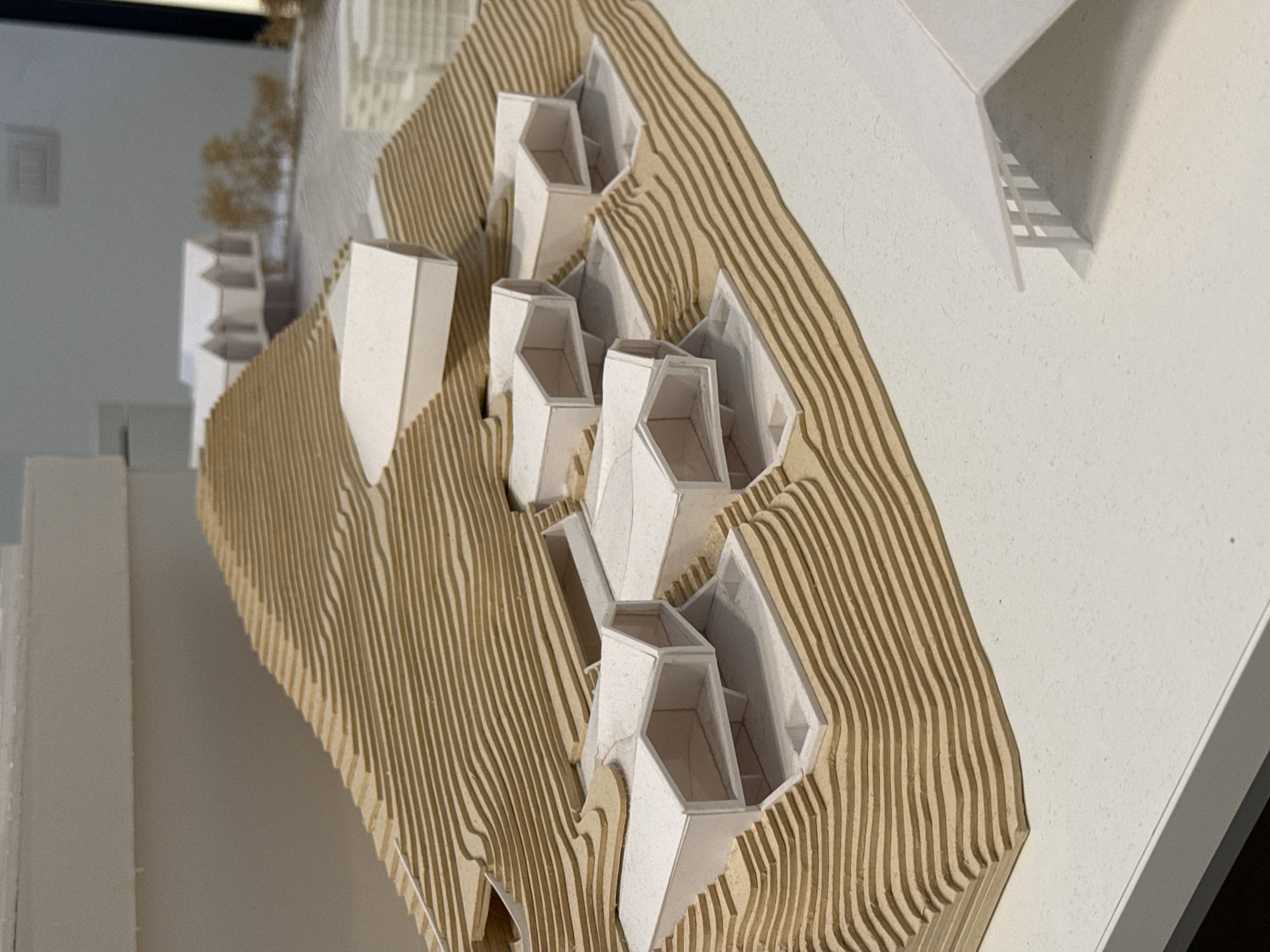 The image presents an architectural model showcasing a proposed development integrated into a contoured landscape. The primary subjects are the model's terrain and the buildings. The terrain is meticulously crafted from numerous horizontal layers of light brown material, likely laser-cut wood or cardboard, forming undulating curves and terraced elevations, indicative of a hilly or sloped site.
Nestled within these contours are several white, angular building structures. These buildings vary slightly in form but share a modern, geometric aesthetic with multiple facets. Many of them feature open upper sections, revealing simplified interior spaces that include miniature stairs or floor levels, suggesting multi-story residential units or individual structures. The buildings are arranged to follow the topography, appearing to step down or rise with the landscape.
In the background, out of focus, are additional elements of the model, including abstract, brownish, textured forms resembling miniature trees, and other white building blocks. A larger, abstract white structure in the upper right casts distinct shadows, revealing what appears to be a slatted or stepped underside. The model is placed on a smooth, light-colored surface, possibly a display table. The lighting, possibly artificial, casts clear shadows that highlight the three-dimensional quality and intricate details of the model.
The overall scene depicts an architectural concept, likely for a hillside or terraced development, emphasizing the relationship between built form and natural or sculpted terrain. The precise construction and clean aesthetic suggest a professional architectural presentation. No discernible text is visible within the image. Given the context, this model could be part of an architectural design project or exhibition in Milan, Italy.
