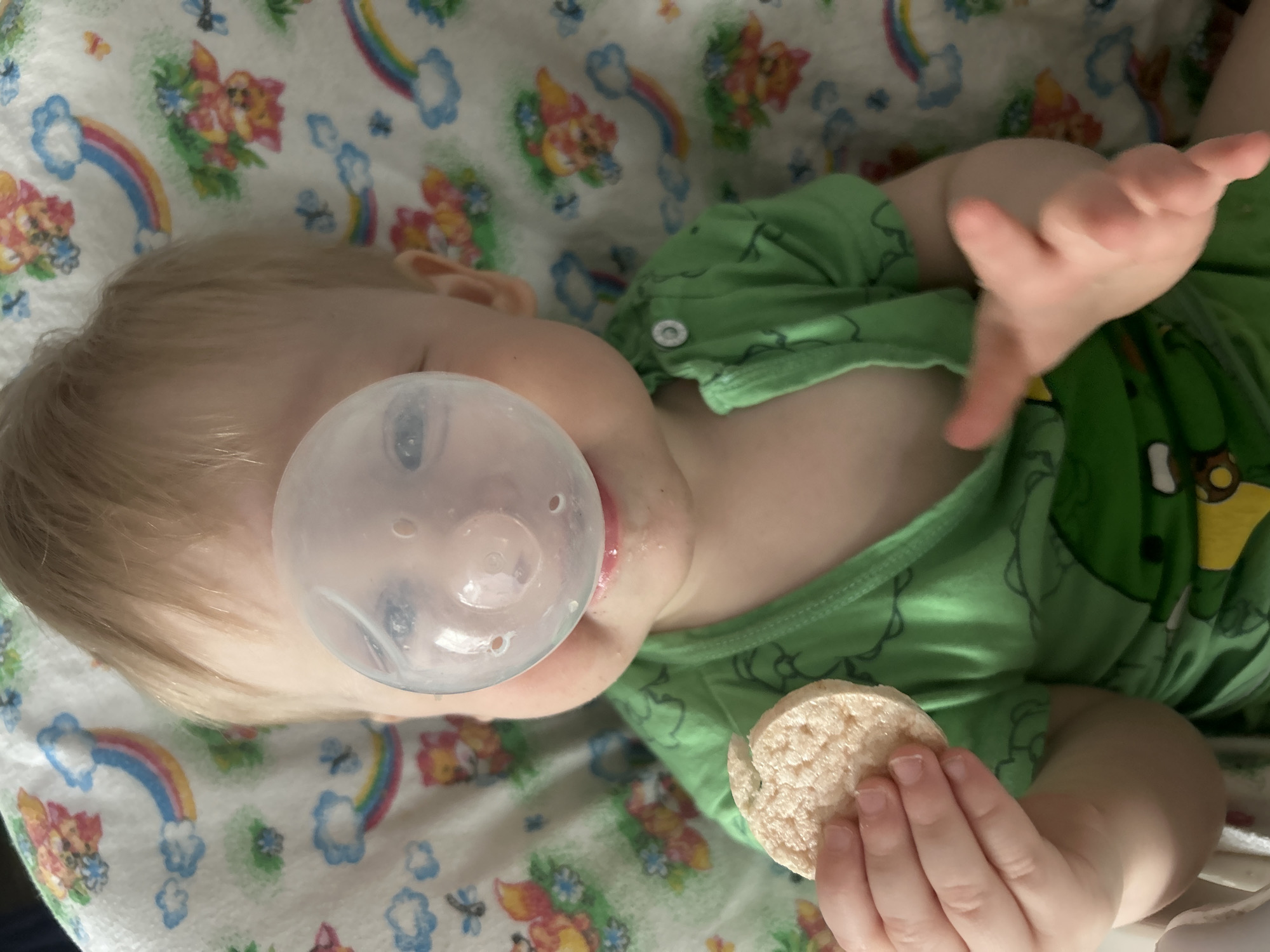 A baby lies on a bed or changing pad patterned with rainbows, flowers, and cartoon bears. The baby is wearing a green shirt, has fair skin, and is using a pacifier. In one hand, the baby holds a small, round rice cake. The location is not discernible from the image.