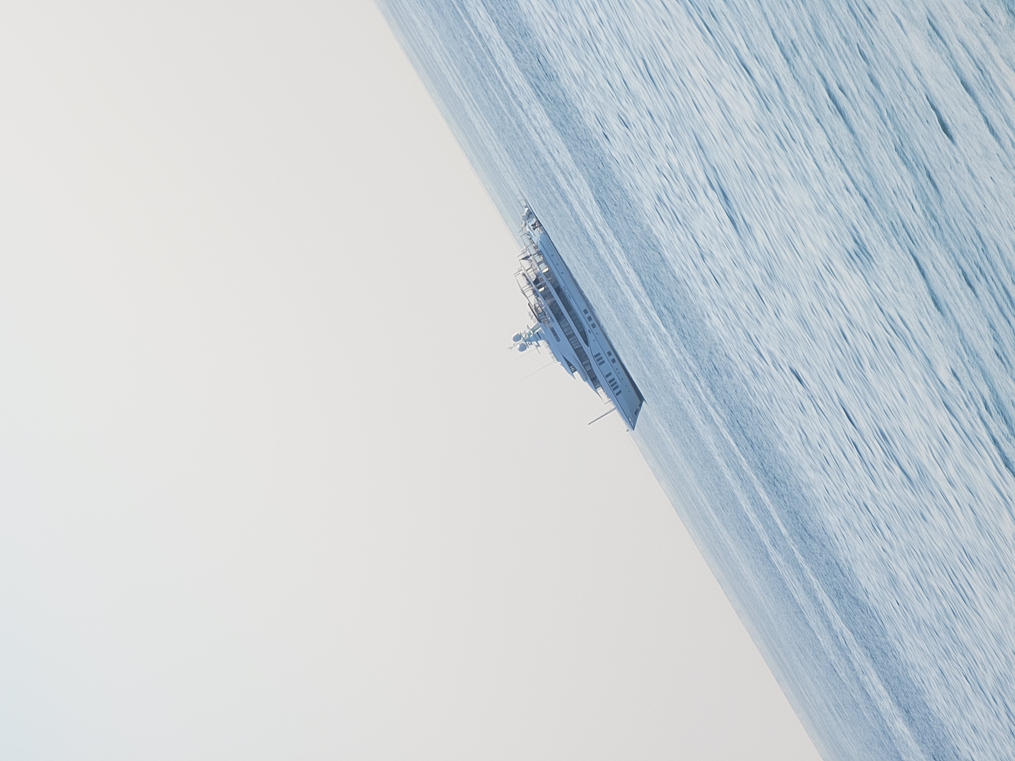 A large, white yacht is docked on a calm, light blue ocean. The horizon is visible at an angle, with the sky a pale, overcast white. The yacht is positioned in the middle right of the frame, at a slight diagonal. It has multiple decks and appears to be luxurious. The water has a gentle ripple pattern. The image is minimalist and serene, with the yacht being the sole prominent subject against the expansive sea and sky. The location is offshore Quarteira, Portugal.