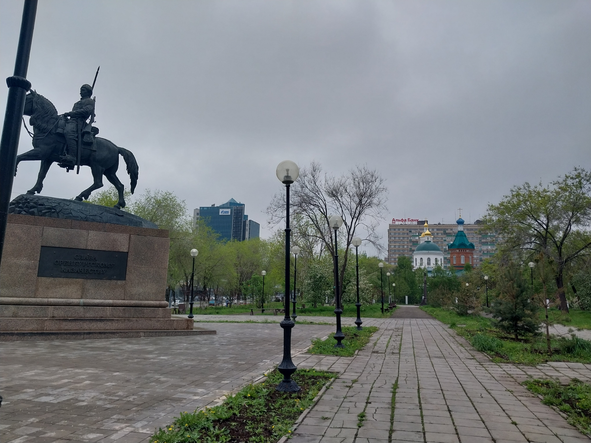 The image depicts a park in Orenburg, Russia, under an overcast sky. In the foreground stands a bronze statue of a man on horseback atop a stone pedestal. The inscription on the pedestal reads "Слава Оренбургскому Казачеству" (Glory to the Orenburg Cossacks). A paved walkway leads into the distance, flanked by ornate black lampposts and patches of grass with small yellow flowers. In the background, buildings are visible, including a modern glass skyscraper and a building with the sign "Альфа Банк" (Alfa Bank). Also in the background is the colorful Church of the Kazan Icon of the Mother of God, recognizable by its green and blue domes and red brickwork. The park appears to be empty of people at the time the photograph was taken.
