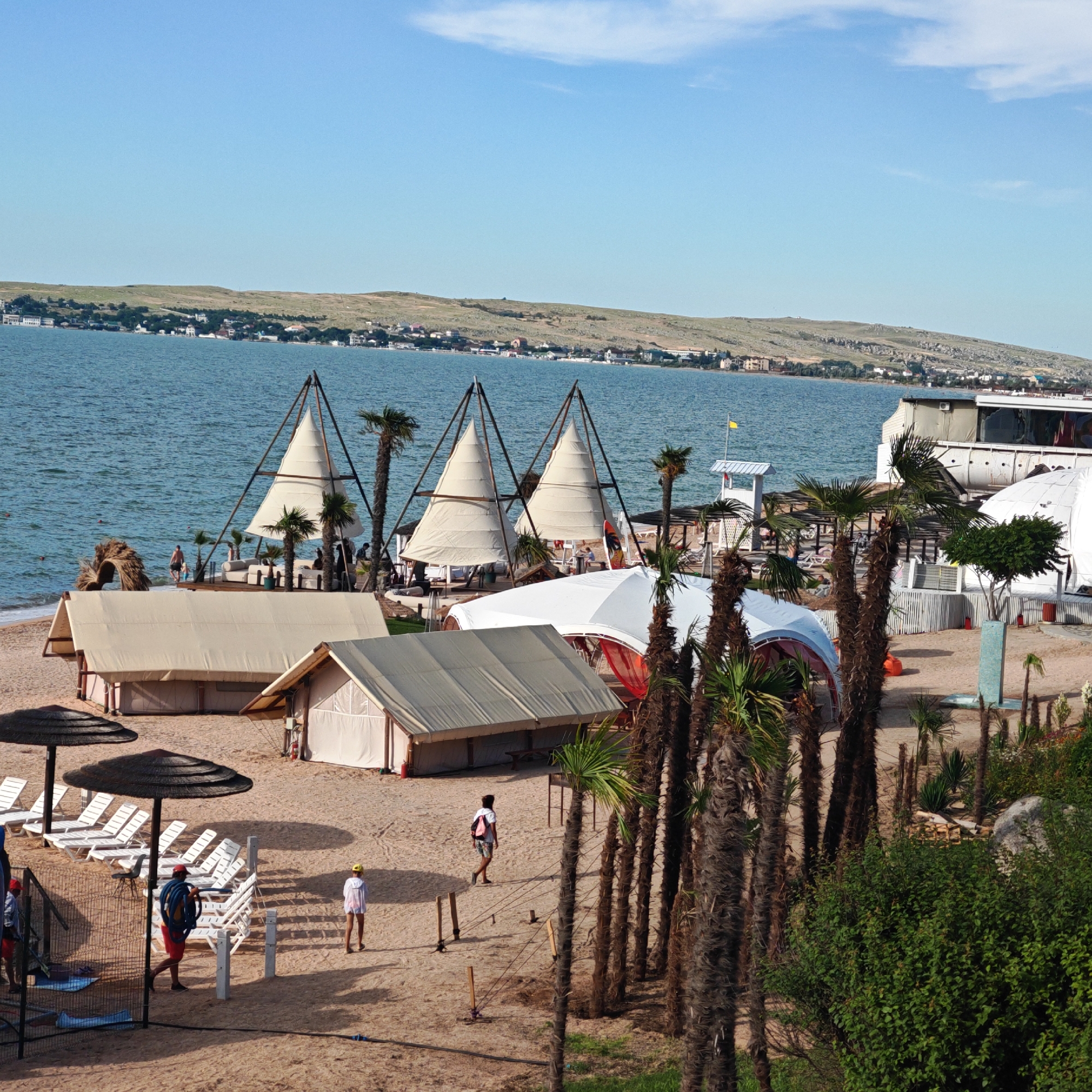 The image depicts a beach resort scene with a body of water in the background, likely a sea or large lake, and a coastline with buildings and vegetation visible on a distant hill. The foreground is a sandy beach with several structures and people.
Prominently featured are several large, tent-like structures with white fabric coverings, suggesting temporary accommodations or event spaces. Some of these structures have triangular designs, supported by metal frames, resembling sails. Adjacent to these are smaller, more traditional tent structures.
Scattered across the beach are white lounge chairs, some arranged in rows under dark, conical beach umbrellas. A few palm trees are present, adding to the tropical atmosphere.
There are several individuals visible in the image. One man in red shorts and a red cap is carrying a coiled blue hose, suggesting he might be a worker attending to the facilities. Another person, a woman in a white hooded jacket and shorts, walks away from the viewer. Further in the background, near the water's edge, a man in dark shorts is walking into the water, accompanied by another person. Other figures are partially visible in the distance, some near the lounge chairs.
In the background, a white building with a dark roof and windows is situated on the beach. A lifeguard tower with a yellow flag is also visible. The sea is a tranquil blue, with gentle waves lapping at the shore. The sky is clear with a few wispy clouds. The overall impression is a well-maintained beach resort, possibly during a summer day.