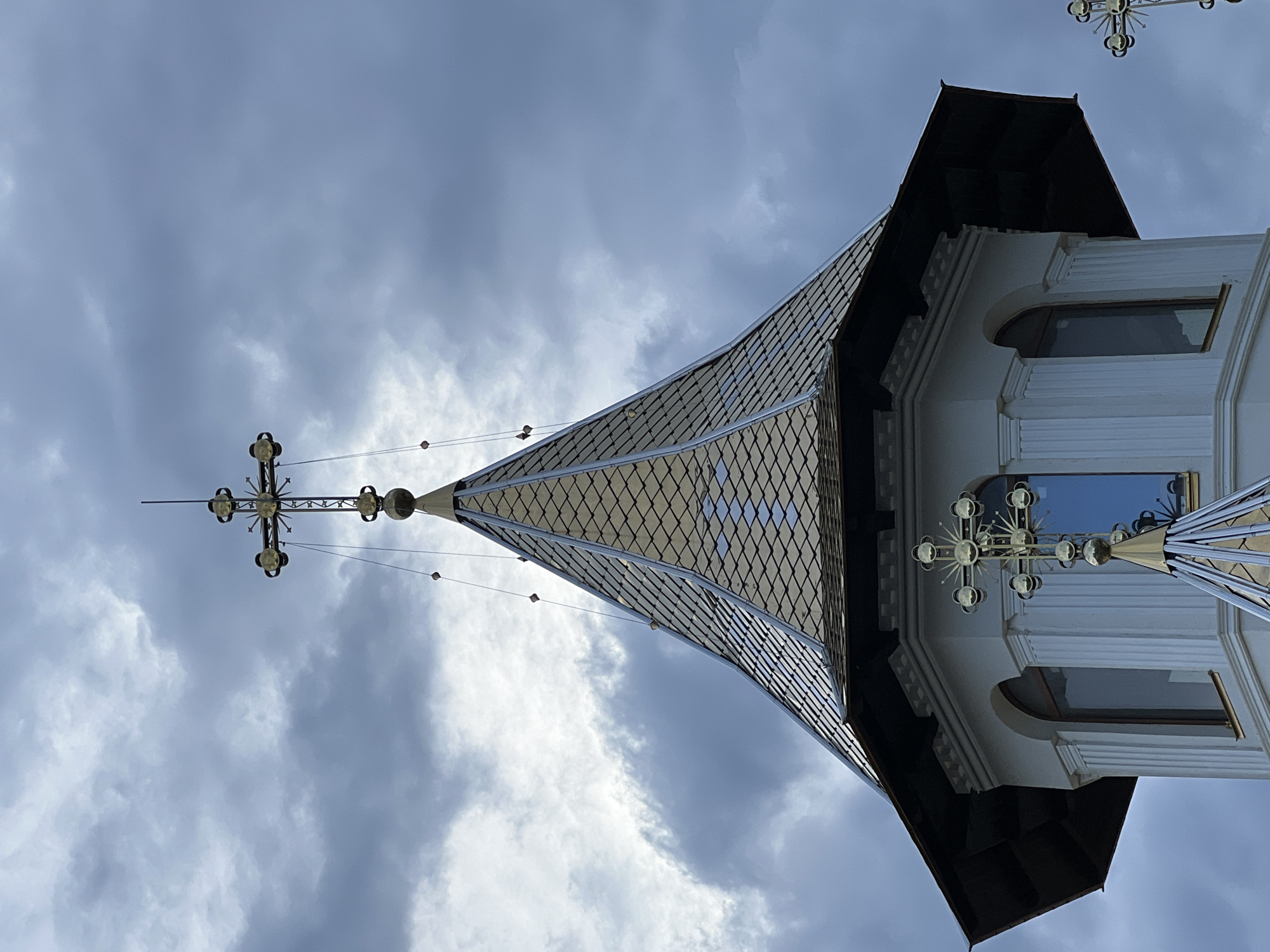 The image shows the upper section of a white church tower with a gold-colored, pointed roof in Năvodari, Romania. The roof is covered in diamond-shaped tiles. A large gold cross with decorative elements sits atop the roof, with thinner wires extending downwards from it. Another, smaller gold cross is visible lower down on the tower, positioned above a window. The tower has arched windows and fluted columns. The sky is overcast with dark, dramatic clouds. The overall scene is serene and spiritual.