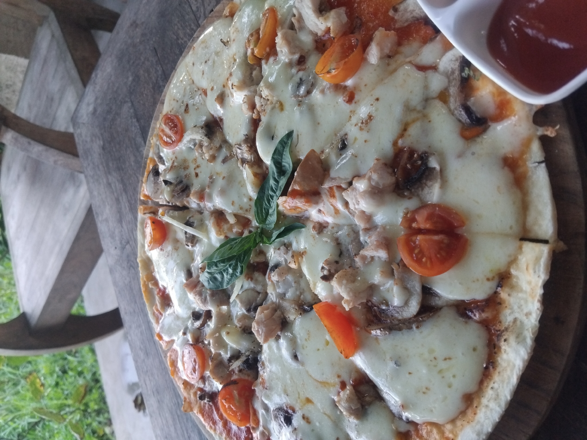 The image shows a pizza with melted cheese, mushrooms, chicken pieces, and tomato slices. A small dish containing a red sauce sits to the right of the pizza. A few basil leaves are placed in the center. The pizza sits on a wooden surface and is near a light gray wooden chair. In the background, green vegetation is visible. The location is Unknown, Unknown.