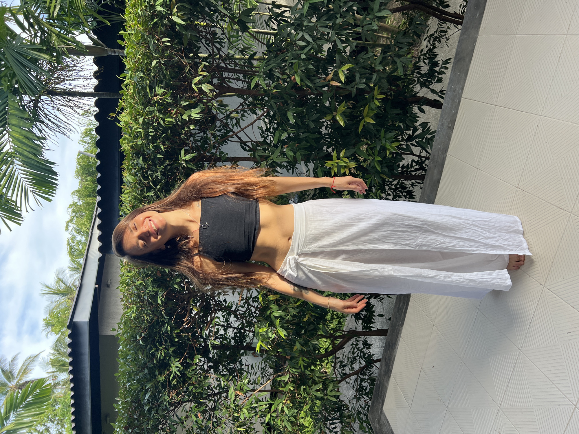 The image depicts a young woman standing on a white tiled surface outdoors. She is wearing a black crop top and long white pants. The woman is positioned in front of a dense green hedge or foliage. Behind the hedge, a structure with a black roof and tropical trees are visible, which suggests that the location is in บ้านบางรักษ์, Thailand. The woman is smiling, and her hair is loose. The setting appears to be a sunny, tropical environment.