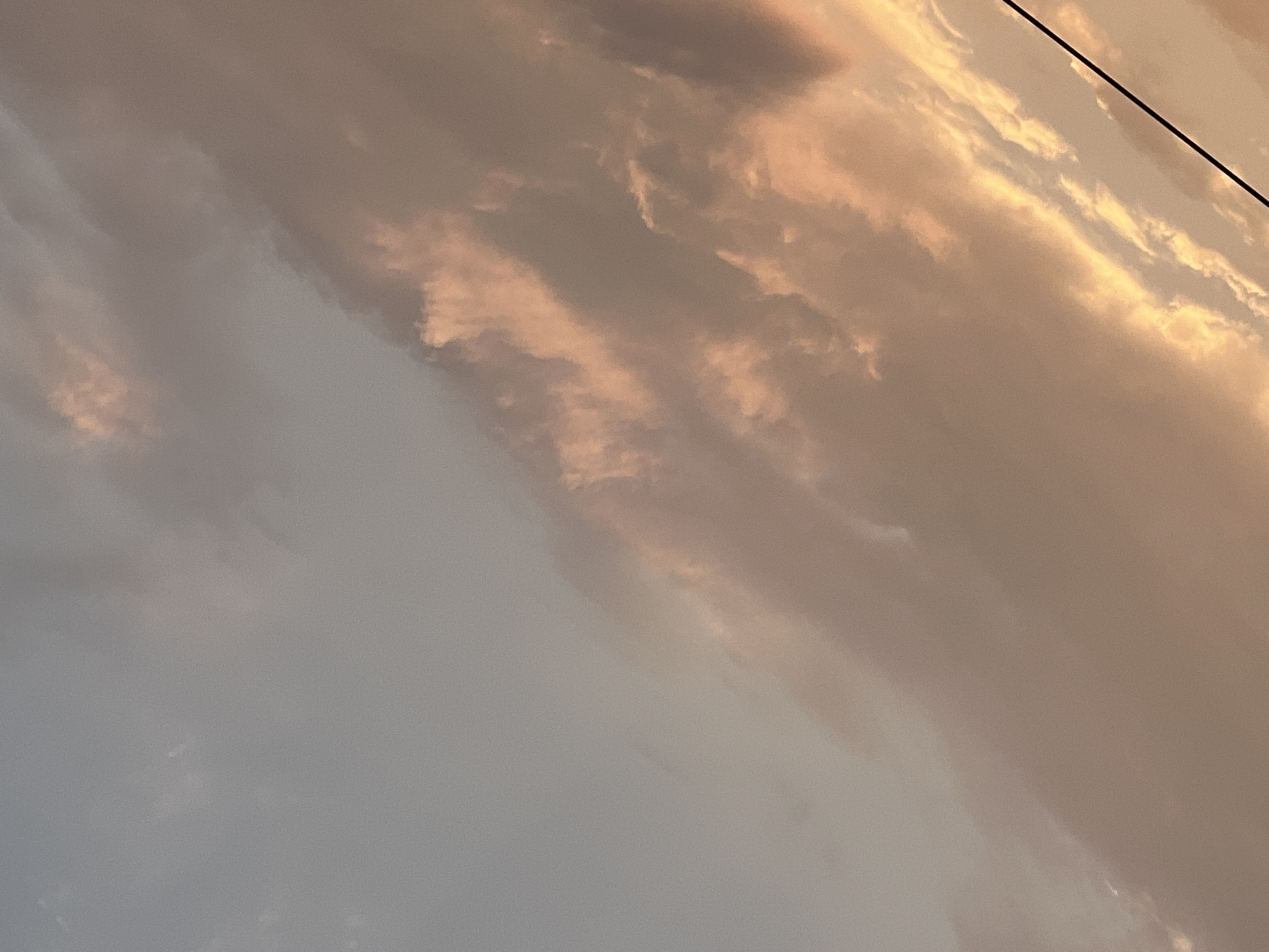 The image depicts a sky with clouds tinged with pink and orange hues, suggesting either sunrise or sunset. The lighting is soft and diffuse. In the lower right quadrant, a black wire, possibly a power line or telephone wire, runs diagonally across the frame. The background is an expansive sky with varying cloud formations, some appearing wispy and others more dense. The scene is devoid of any discernable landmarks that would pinpoint its location within Warsaw, Poland. There are no people, animals, or specific objects in the foreground or middle ground that would provide further context or details about the environment beyond the sky and the wire. The overall impression is one of a natural, atmospheric phenomenon.