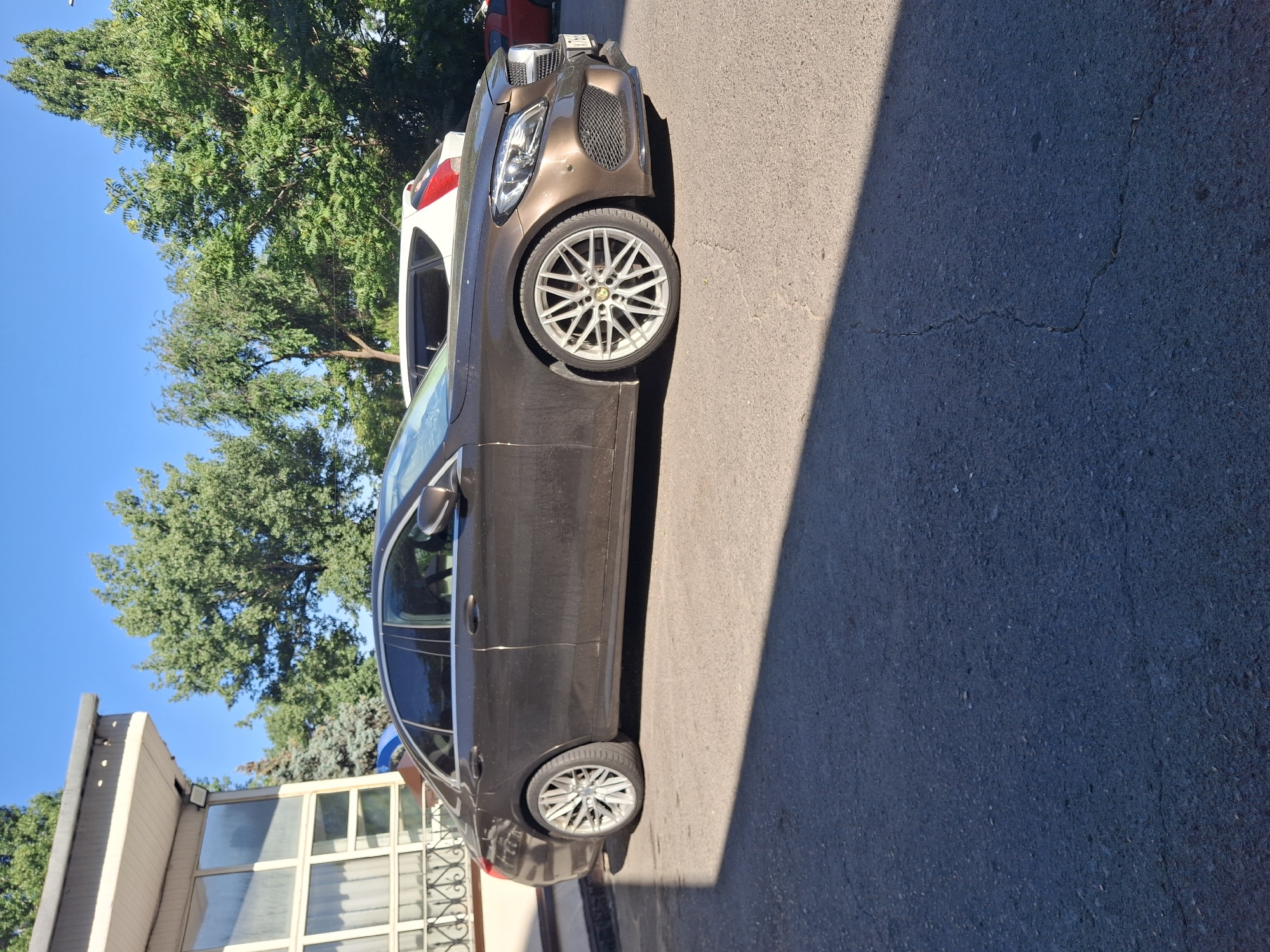 The media file shows a brown car parked on an asphalt surface, casting a shadow to the right. The car is positioned sideways, with its front on the left and rear on the right. It has a sleek design with alloy wheels. Behind the car, there is a white SUV and a red car, partially visible.
In the background, there are trees with green foliage and a clear blue sky, suggesting a sunny day. To the left of the car, there is a building with large windows, indicating an urban or suburban setting. The building appears to be a commercial or residential structure.
The overall scene depicts a parking area on a bright day, likely in Rostov-on-Don, Russia, given the location context. There is no activity taking place, as the cars are parked. The sunlight and shadows suggest it is daytime. No people are visible in the image. The asphalt ground shows some cracks. There is no visible text in the image.