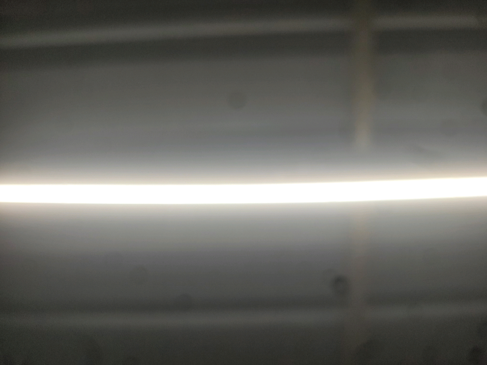 The image shows a blurred, horizontal bright light source against a dark, textured background. The light appears to be a fluorescent tube or LED strip, emitting a strong, even glow. The background is a gradient of gray tones, with some faint horizontal lines or shadows visible, suggesting a ceiling or an interior surface. There are also a few indistinct dark spots scattered across the background. The overall impression is of an indoor environment, possibly a corridor or a room with artificial lighting. The specific location of Santekatte, India, is not discernible from the visual information provided.