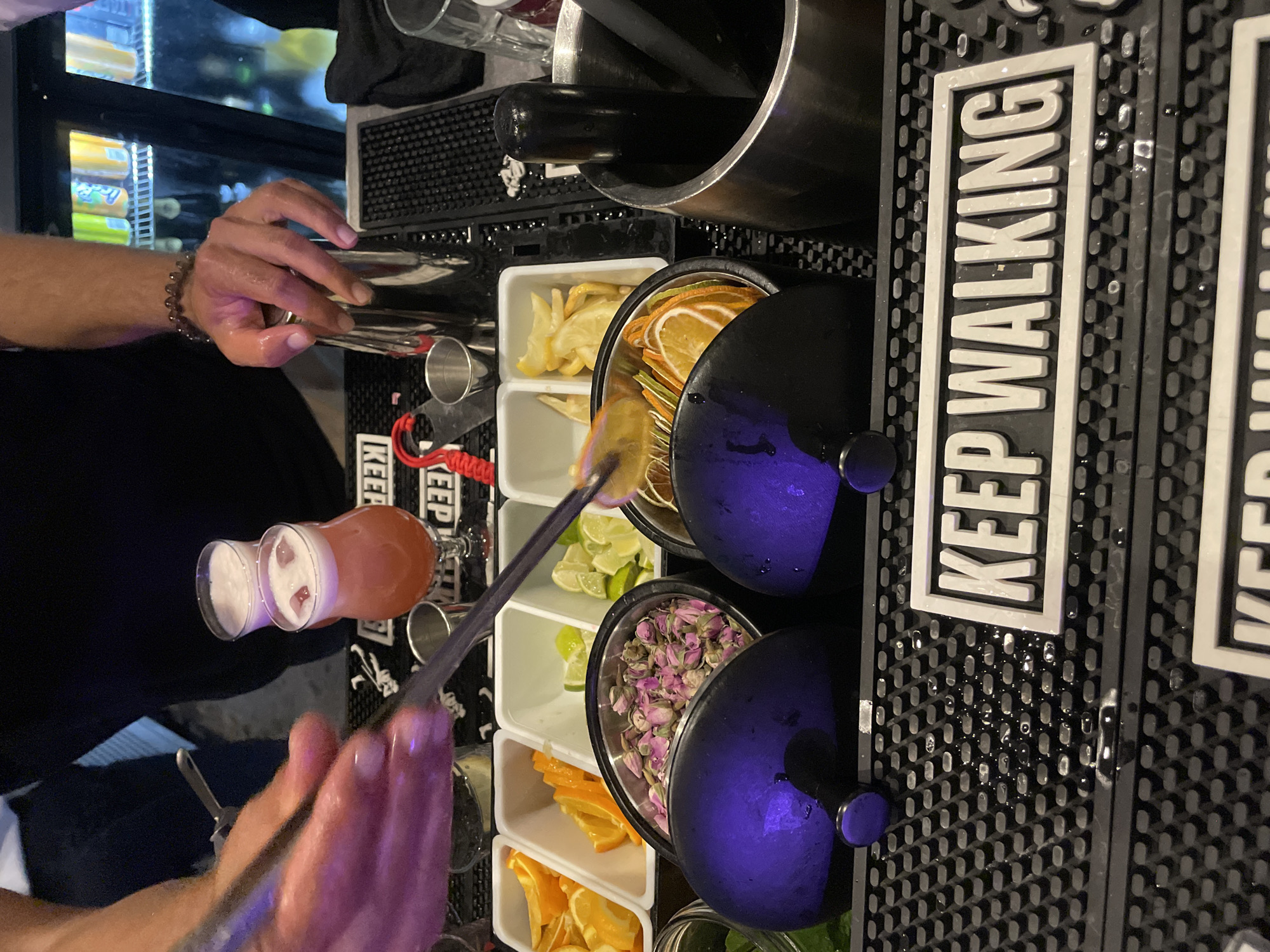 The image captures a close-up view of a bartender's hands preparing a drink at a bar. The setting appears to be indoors, possibly a cocktail bar, with a blurred background showing shelves stocked with beverages, including what appear to be colorful soda cans. In the foreground, a black bar mat is visible, with the white lettering "KEEP WALKING" embossed on it.
The bartender's hands are the central focus. One hand, wearing a bracelet, is gripping a silver cocktail shaker. The other hand, with fingers extended, is holding a metal utensil, seemingly to add garnishes to a drink.
On the bar surface, several small white rectangular containers hold various drink garnishes. These include sliced oranges, sliced limes, dried citrus slices, and what appear to be dried rosebuds. Two small cocktail glasses are also present; one contains a pinkish liquid topped with foam and ice, and the other is partially obscured but also appears to contain a frothy drink.
A large, dark metal mortar and pestle are positioned on the right side of the frame, with a black pestle inside. Two dark purple, bowl-like objects with knobs are in the lower portion of the image, casting a violet glow. The bar mat is wet, with visible droplets of liquid scattered across its surface.
The image does not contain any information or visual cues to indicate its location in the city of Obzor, Bulgaria.