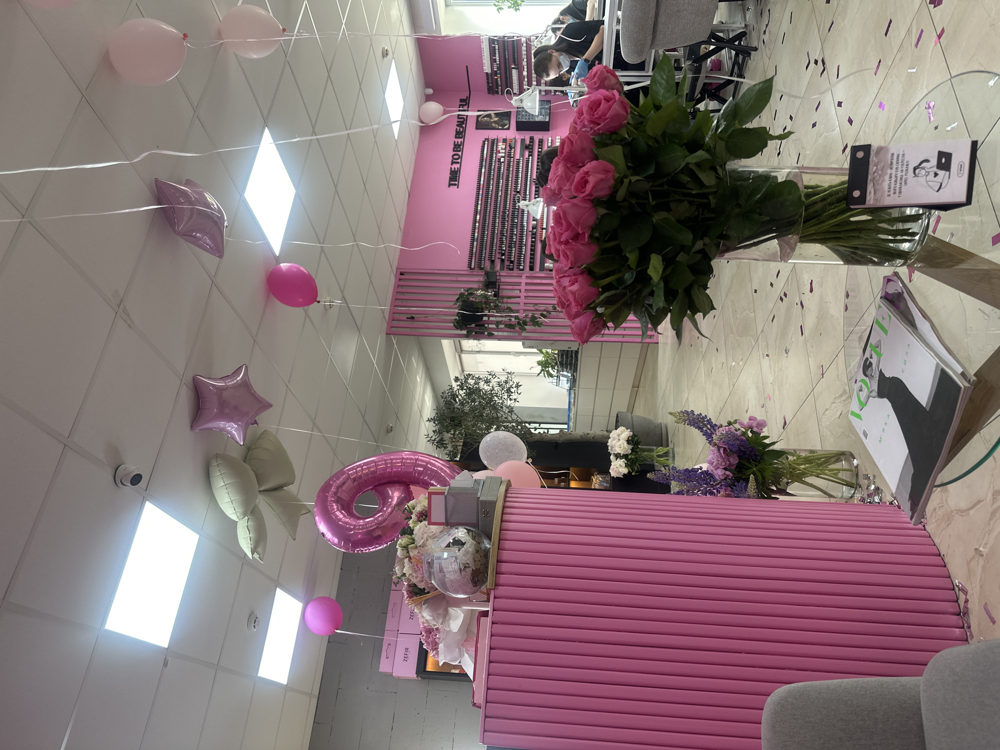 This media file captures a festive and brightly lit beauty salon in Ternopil, Ukraine, decorated for a special occasion. The predominant color theme is pink, evident in the large bouquet of pink roses on a glass table, the ribbed reception desk, wall panels, and numerous balloons.
The ceiling is adorned with a string of pink and light-colored balloons, including star-shaped foil balloons and one large foil balloon shaped like the number '6', suggesting a 6th anniversary or birthday celebration. Confetti is scattered across the tiled floor, further indicating a festive event.
In the background, two individuals are visible, seated and likely providing nail services, indicating the salon is operational despite the celebration. A wall display behind them showcases numerous nail polish bottles. Above this display, the text "TIME TO BE BEAUTIFUL" is prominently featured in white lettering against a pink wall.
The pink reception desk is also decorated with a smaller bouquet of flowers, balloons, and boxes labeled "ZEFIR". On the glass table, next to the large rose bouquet, there's a small stand with Ukrainian text, part of which reads "ДЕ ЛЕЖАТЬ КЛЮЧІ НЕПАЛИТАЯ ЯКОГО КОЛЬОРУ БУВ САР... НА ВЕРЕСНЯ - ЗАРАЗ РОЗКАЖУ" (Where are the unburnt keys What color was the tsar... For September - now I'll tell you). A magazine with the word "СИЛА" (Strength/Power) in Ukrainian is also on the table.
The salon has bright overhead lighting and a security camera mounted on the ceiling. The overall atmosphere is cheerful and welcoming, combining daily salon operations with a celebratory mood.