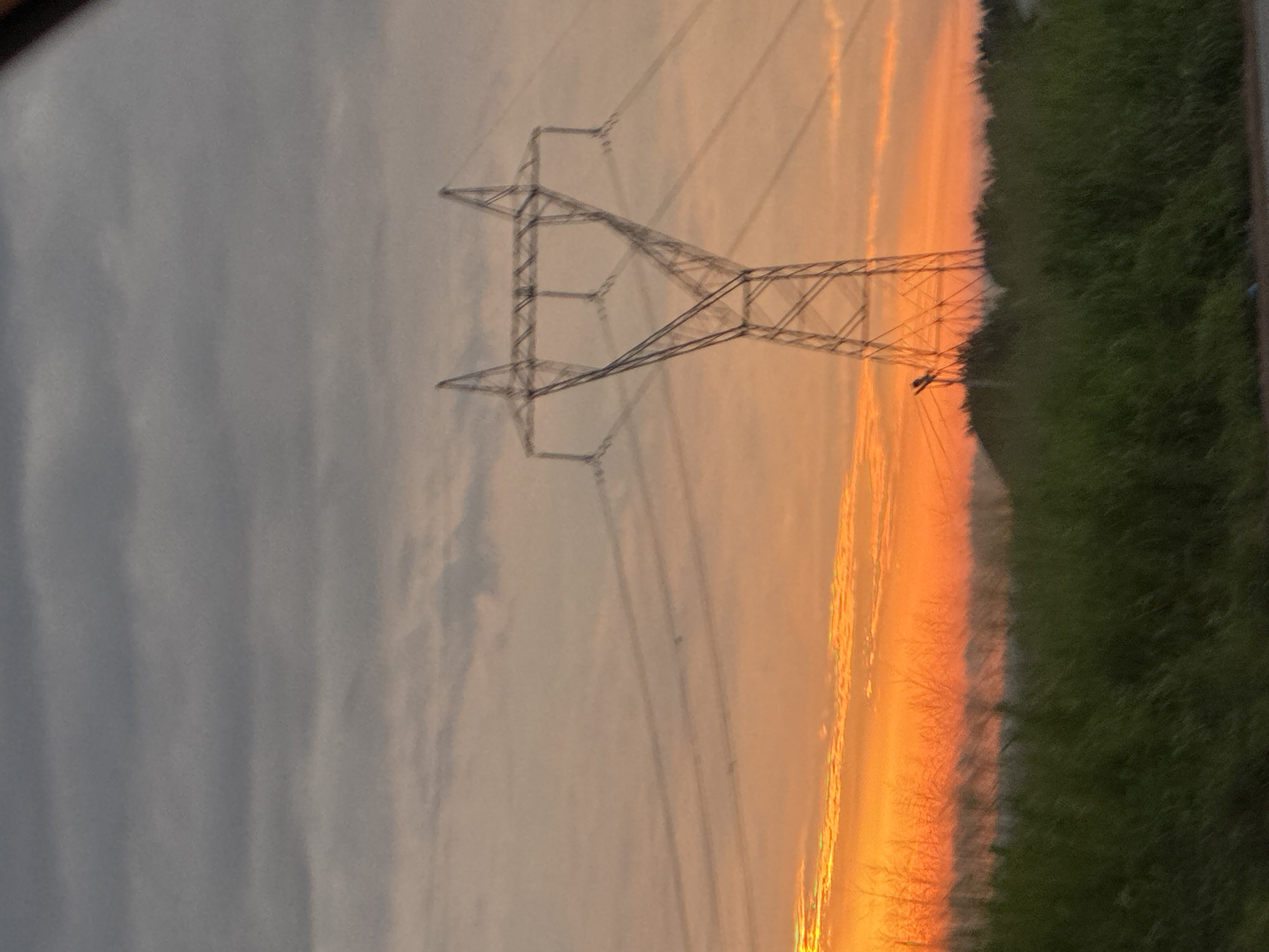 This image depicts a wide shot of a rural landscape at sunset, with a large electricity pylon dominating the center-right of the frame. The sky is a vibrant gradient of orange and yellow near the horizon, transitioning to grey and dark clouds overhead. Several power lines extend from the pylon across the sky. In the foreground, a dense field of green grass or reeds is visible, with hints of darker vegetation and possibly trees in the middle ground. The overall impression is one of a vast, open space illuminated by the dramatic colors of a setting sun. There are no people or identifiable structures in the immediate vicinity of the pylon. The image's quality is slightly blurred, suggesting it may have been taken from a moving vehicle or with an unsteady hand. This scene is likely located in a rural area of Orifite, Nigeria, given the presence of such infrastructure and the natural vegetation.