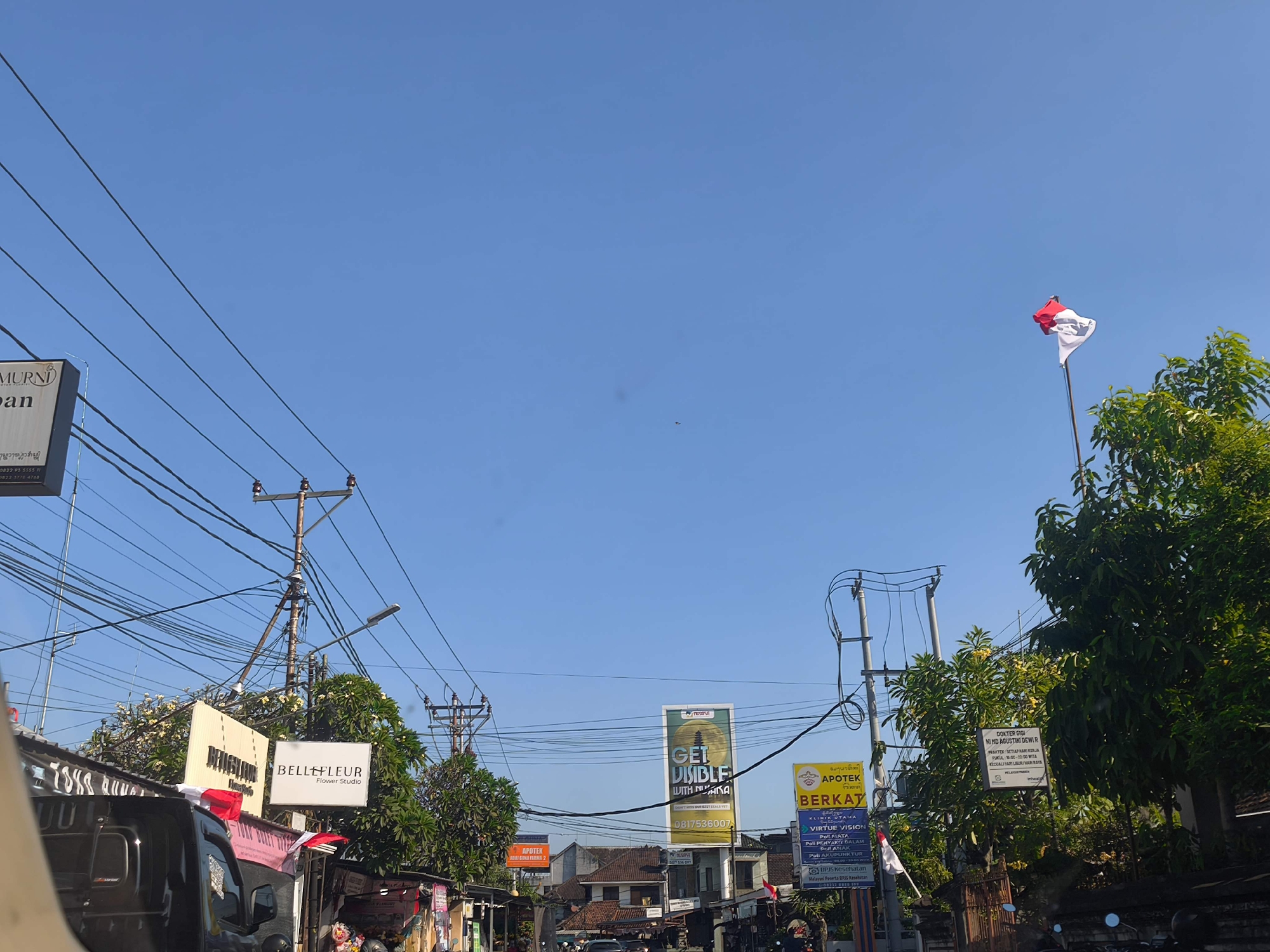 The image shows a street scene in Denpasar, Indonesia, under a clear blue sky. Overhead, a dense network of utility wires crisscrosses the frame, supported by wooden utility poles. Several signs are visible along the street. On the left, a sign reads "MURNI" and "Jinan". Further down the street, a sign for "BELLE FLEUR Flower Studio" is prominent. In the distance, a large billboard displays the text "GET VISIBLE WITH NU ANA". Another sign for "APOTEK BERKAT" is also visible, along with medical clinic information. An Indonesian flag, red and white, flies from a flagpole. Lush green trees line parts of the street, and a black truck is partially visible in the foreground. The environment appears to be a commercial area with shops and businesses.