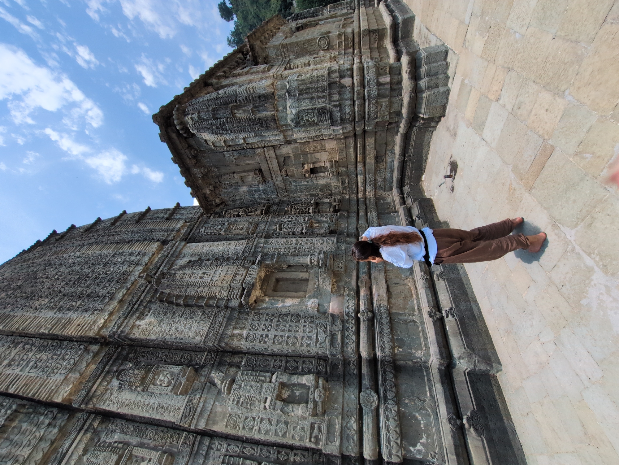 A person stands in front of the intricately carved stone facade of a temple, possibly in Mandi, India, based on its architectural style. The temple is constructed from large, dark grey stone blocks and features elaborate carvings of patterns and figures across its surface. The person, viewed from behind, has long brown hair and is wearing a white long-sleeved shirt and loose brown pants. They appear to be looking up at or interacting with the temple wall. The ground is paved with light-colored flagstones. The sky above is blue with scattered white clouds, and a green hillside is visible in the background.