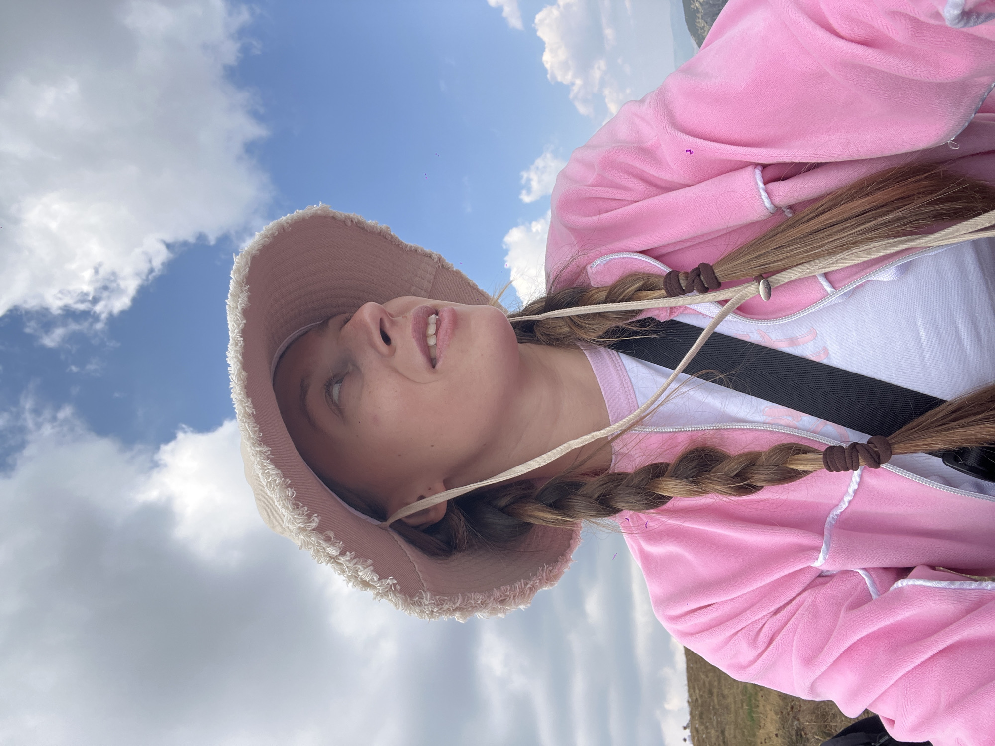A young woman, wearing a pink velour jacket over a white t-shirt and a light-colored bucket hat, looks upwards and to her right. Her hair is styled in two braids, secured with brown ties. She is outdoors, with a cloudy blue sky overhead and a grassy hillside in the background. The location is the town of Боричје, Montenegro.