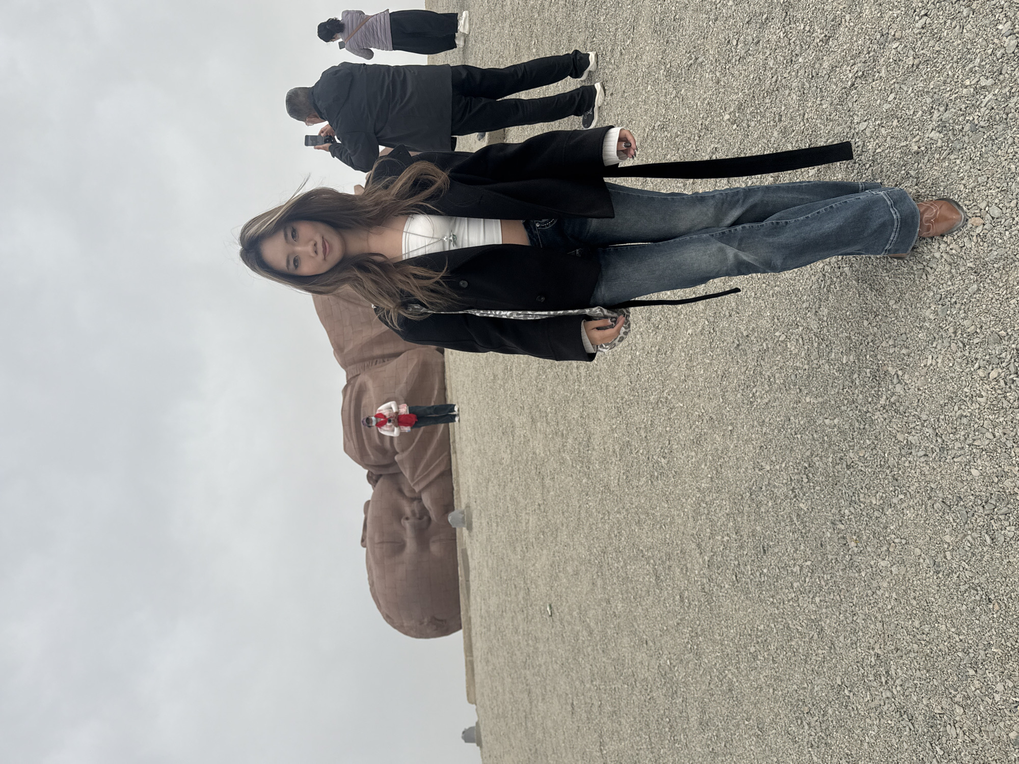The image shows a woman standing near a large, recumbent Buddha statue made of stone, located in Guazhou County, China. The foreground of the image features a woman in jeans, a white top, and a black jacket. The woman stands on a gravel surface. Behind her is the large stone Buddha statue. Several other people are present in the image, including one taking a photograph and another with a purple top. The sky is overcast.