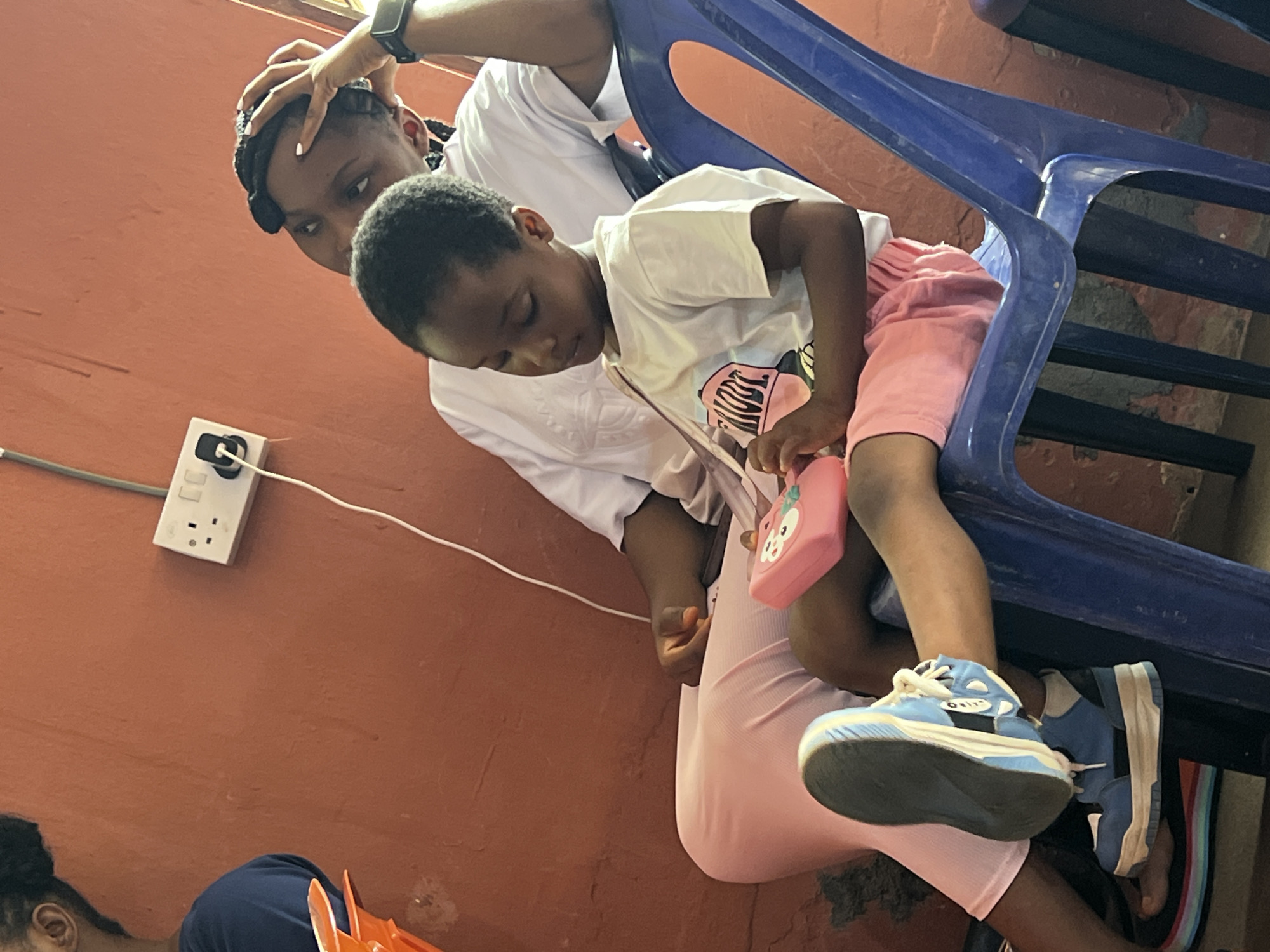 In Nnewi, Nigeria, a young child with dark hair and eyes, wearing a white t-shirt and pink shorts, sits on a blue plastic chair. They are holding a pink toy purse with a cartoon character on it. The child's right leg is crossed over their left, and they are wearing blue and white sneakers. Behind the child, a woman with dark braided hair and braids extending down her back, wearing a white t-shirt, rests her head on her hand, looking to the side. She is wearing a black watch on her left wrist. The background is a terracotta-colored wall with a white electrical socket and a white electrical cord running down it. The image appears to have been taken indoors, with the lighting suggesting it could be daytime.