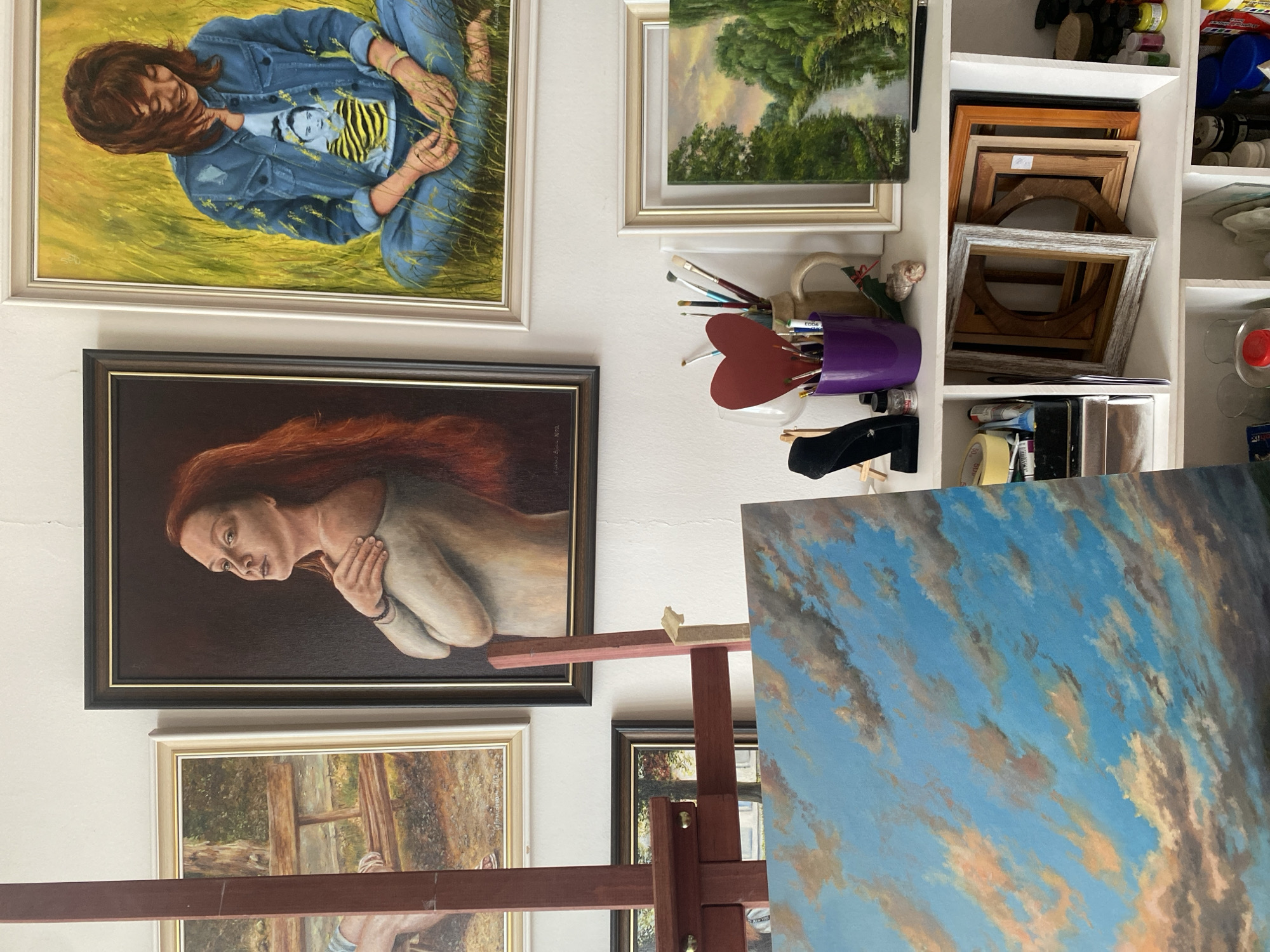 The image depicts an artist's studio, filled with paintings and art supplies. The primary subjects are several framed artworks, including a large portrait of a woman with flowing red hair, a painting of a person in a denim jacket sitting in a grassy field, and a landscape of trees and a river. There are also various art supplies visible, such as paintbrushes in a purple holder, tubes of paint, and rolls of tape. Several empty picture frames are stacked on a shelf. A large canvas depicting a blue sky with clouds is propped up on an easel in the foreground. The setting appears to be an indoor space, likely a home studio, given the amount of personal artwork and supplies. The overall atmosphere suggests a creative and organized workspace. There is no specific activity or event taking place, but the presence of an easel and fresh canvas indicates ongoing artistic creation. No text is clearly legible in the image, beyond what might be on paint labels or other small items.
