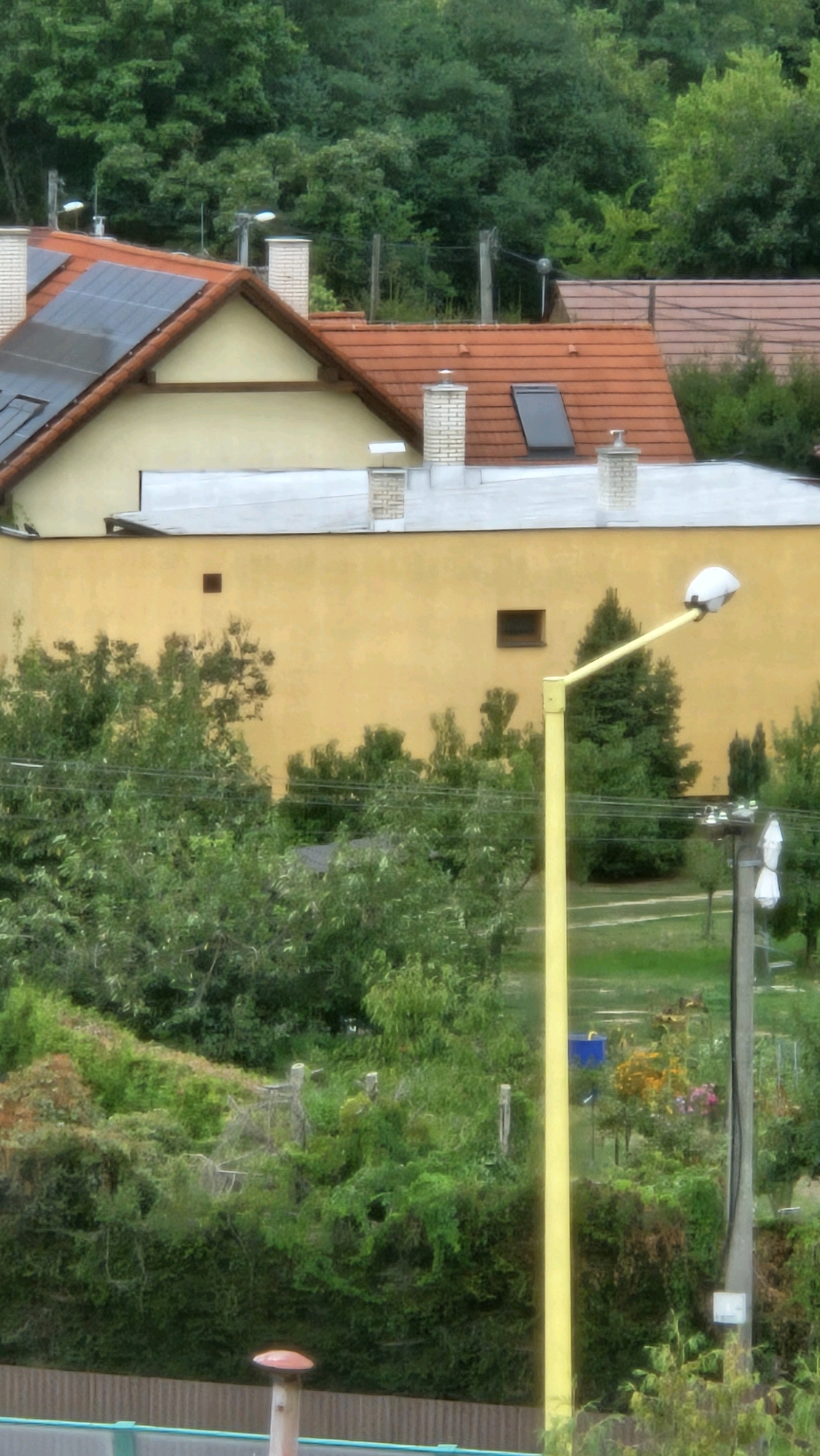 The image shows a street view in Prešov, Slovakia, focusing on a residential building with a yellow facade. The building has a red tiled roof with solar panels and dormer windows. In the foreground, a bright yellow street lamp with a white lamp head extends upwards. Behind the building, dense green trees form a backdrop, with a glimpse of a hill or forest. A concrete utility pole with white insulators and what appear to be several white fixtures is visible to the right of the street lamp. The scene depicts a suburban or semi-rural environment within the city.