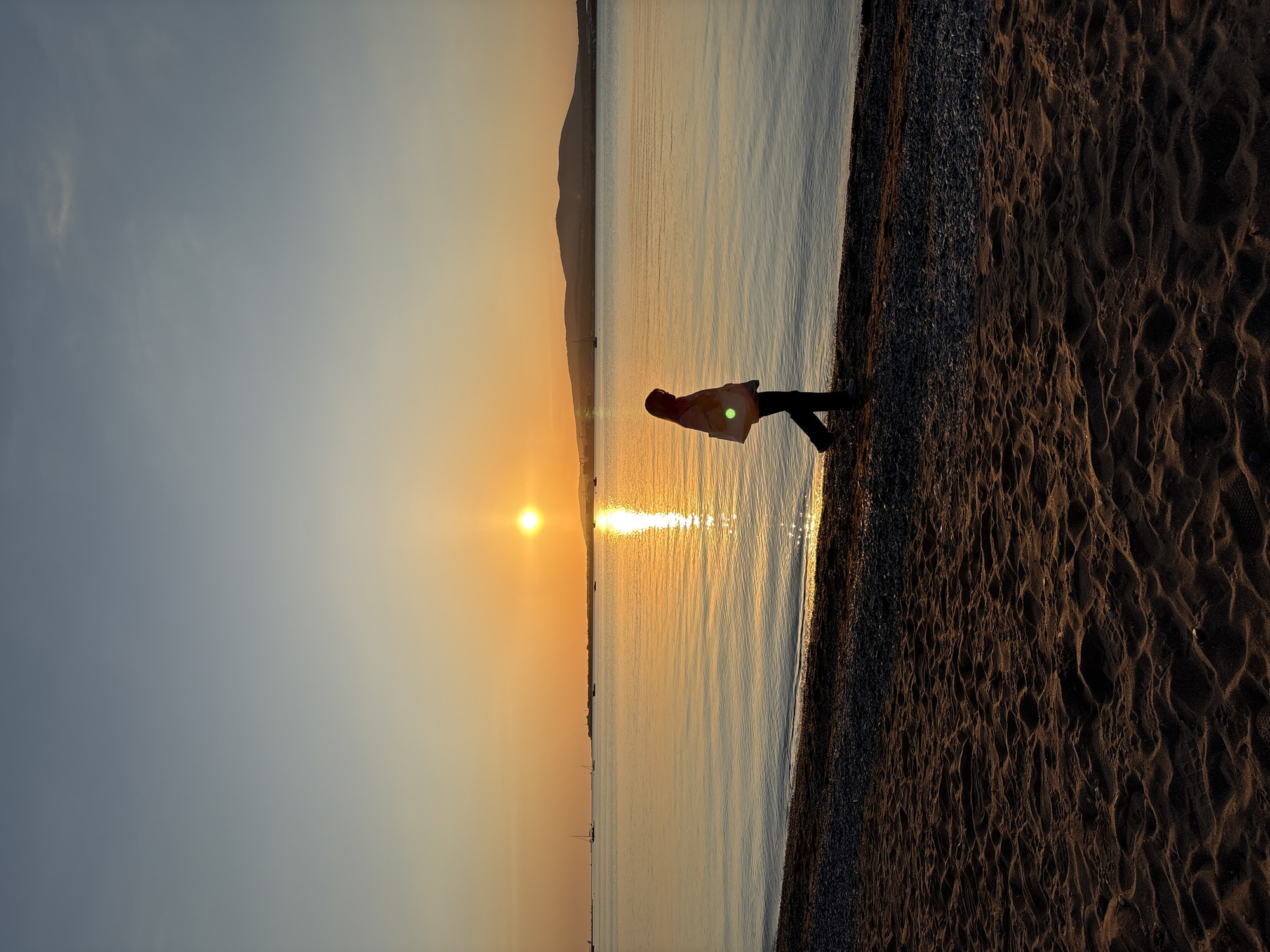The image captures a tranquil coastal scene at sunset in Gelendzhik, Russia. A lone figure stands in silhouette at the water's edge, their back slightly turned towards the camera, observing the vibrant setting sun.
The sun, a brilliant golden orb, is positioned low on the horizon, casting a long, shimmering path of light across the calm water of the sea. The sky above displays a gradient of colors, transitioning from a soft blue-grey at the top to warm orange and yellow tones near the horizon. In the distance, a dark, undulating silhouette of mountains or land stretches across the horizon line. Several small boats are faintly visible on the water, adding to the sense of a populated coast.
The foreground features a textured beach. Closest to the water is a strip of dark pebbles, which then transitions into a wider expanse of dark sand, marked with distinct, undulating patterns and footprints, illuminated by the warm, low light. A subtle green lens flare is noticeable near the person's torso.
The overall atmosphere is peaceful and contemplative, highlighting the serene beauty of the sunset over the sea. The time of day is clearly evening, and the weather appears clear and calm. No text is visible in the image.