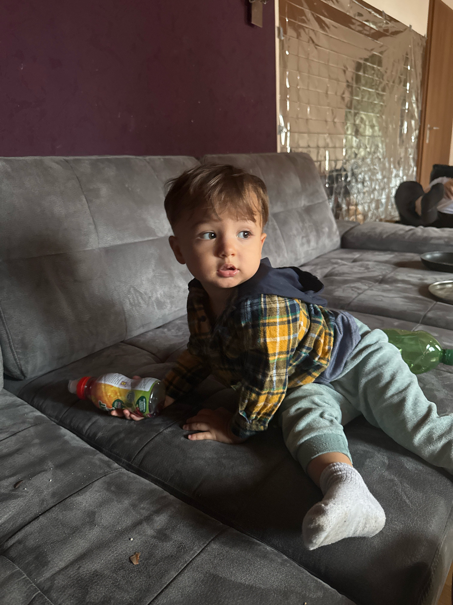A young child is sitting on a gray sofa, holding a bottle of juice. The child is wearing a plaid shirt and light blue pants with a white sock on their right foot. The sofa is in a room with a purple wall and a metallic curtain covering a window. There is a green plastic bottle on the sofa next to the child. The scene appears to be in a domestic setting. The location in Überherrn, Germany is not discernible from the image.