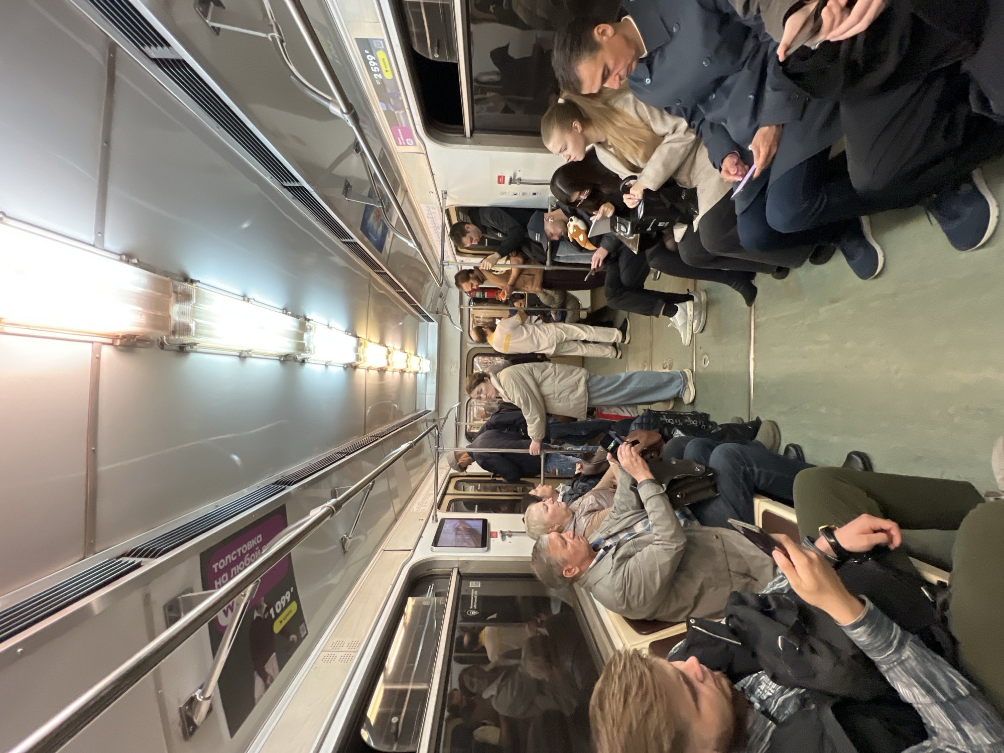 The image shows the interior of a subway car in Moscow, Russia. A significant number of passengers are seated, mostly occupied with their phones. Some passengers are wearing face masks. Advertisements are visible on the walls above the windows and along the side of the car. The lighting inside the subway car is fluorescent.