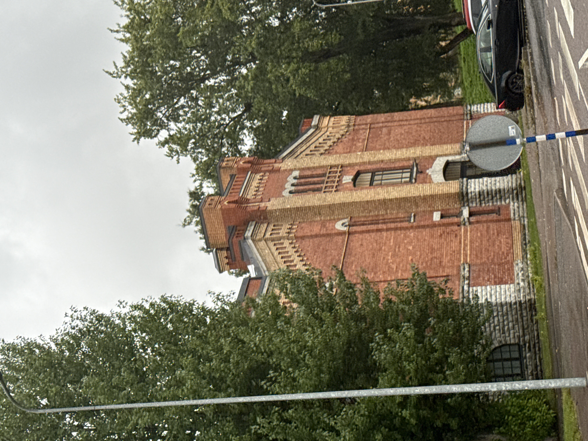 The image depicts a section of a brick building in Tallinn, Estonia, with a prominent streetlamp on the left and lush green trees surrounding the structure. The building's facade is characterized by red brickwork with lighter colored brick accents and stone foundations. An arched doorway is visible at the base of the building. A black car is parked on the right side of the road in front of a grassy incline. The sky is overcast, suggesting a cloudy day.
