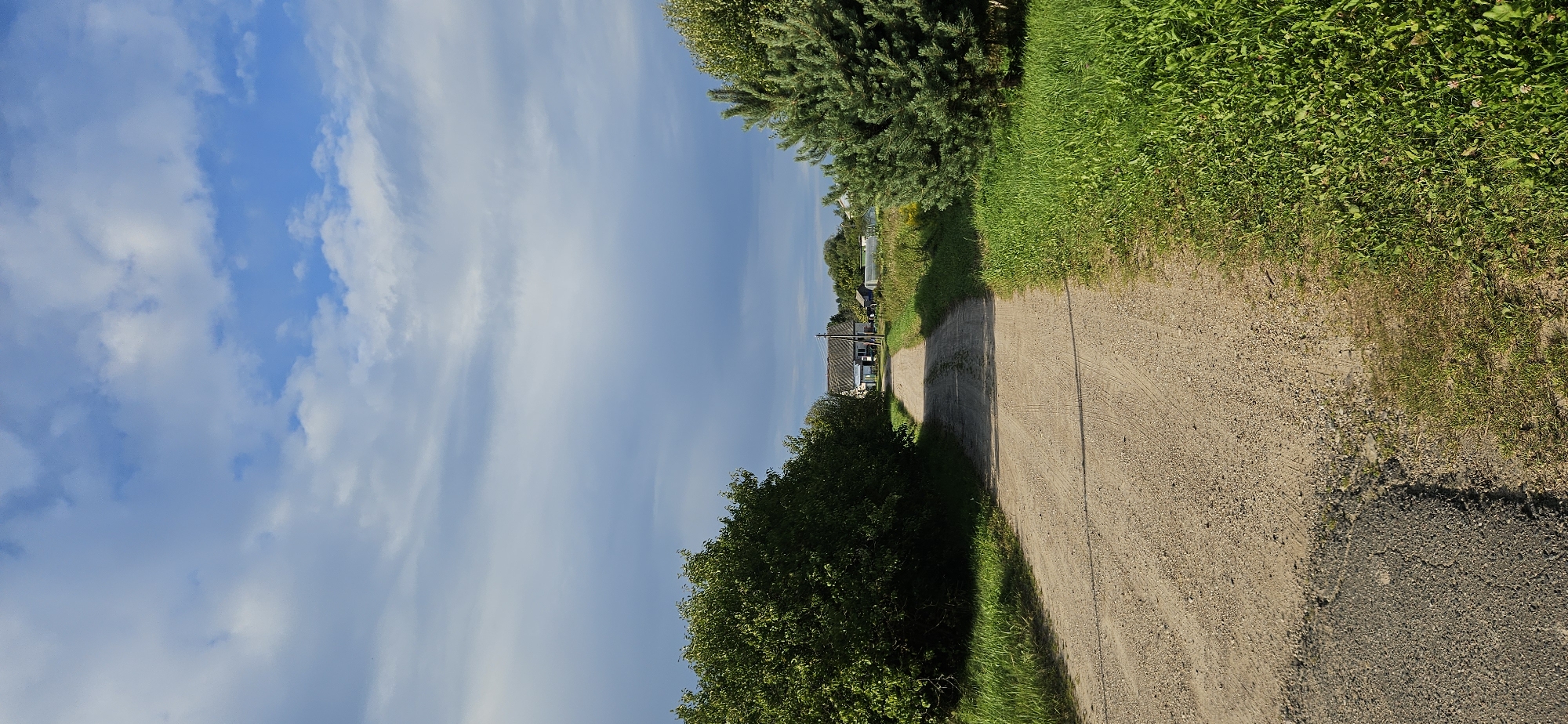 The image shows a dirt road leading towards a cluster of buildings in a rural setting. The road is flanked by lush green grass and trees on either side. In the distance, a few houses and what appear to be greenhouses are visible. The sky is partly cloudy, with blue patches interspersed with white and grey clouds. The scene appears to be a quiet, undeveloped area. No people or animals are present in the image. The location is specified as Мазалаўскі сельскі Савет, Belarus.