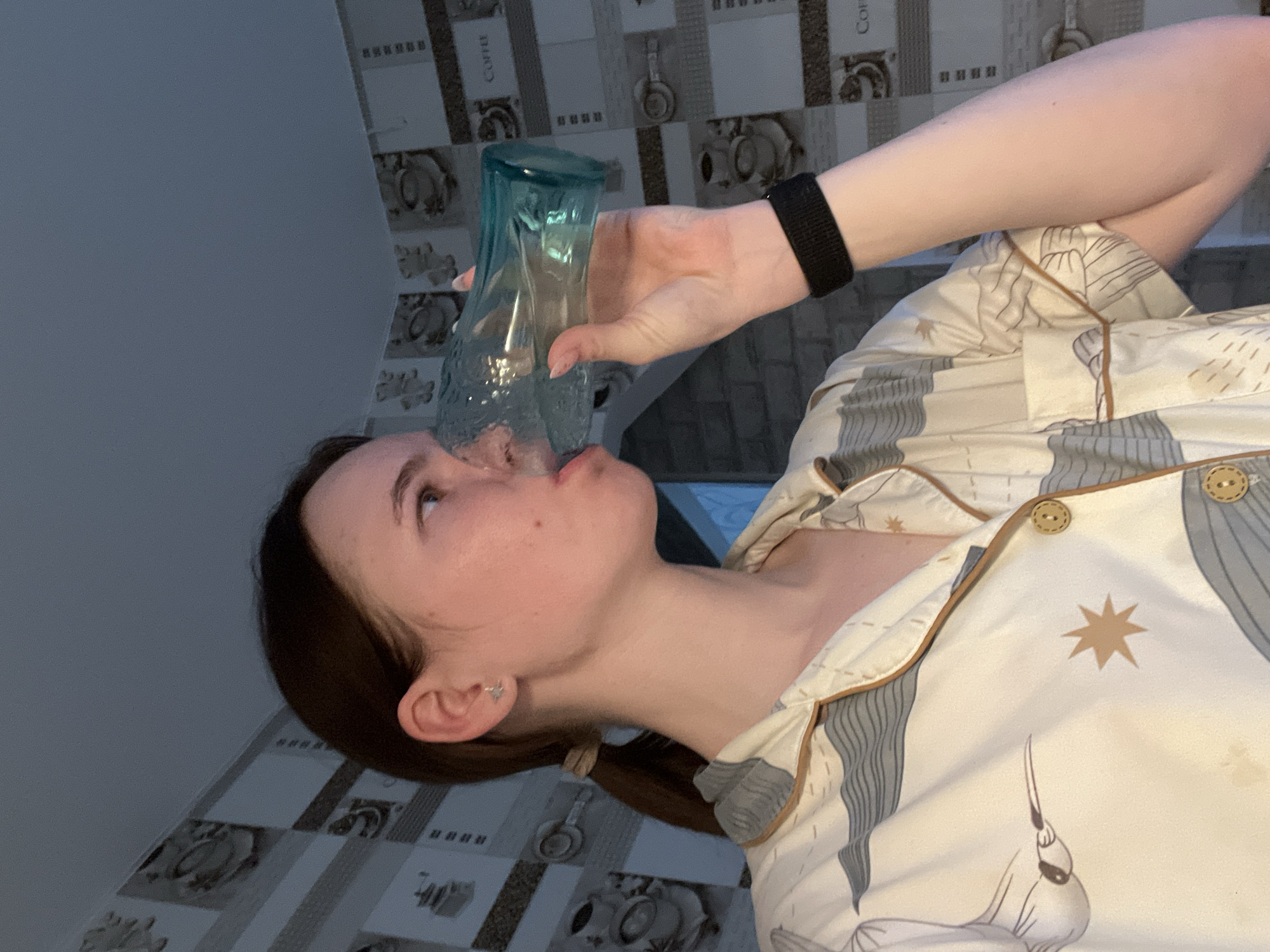 A person is drinking from a light blue, textured glass. The person has brown hair pulled back in a low ponytail, fair skin, and is wearing a star-shaped earring in the left ear. A black fitness tracker is on the person's left wrist. The person is wearing light-colored pajamas with a pattern featuring stars and what appears to be a bird design, outlined in brown piping, and closed with tan-colored buttons. The background consists of tiled walls with a repeating pattern of coffee-related items. The location is Chaikovsky, Russia.