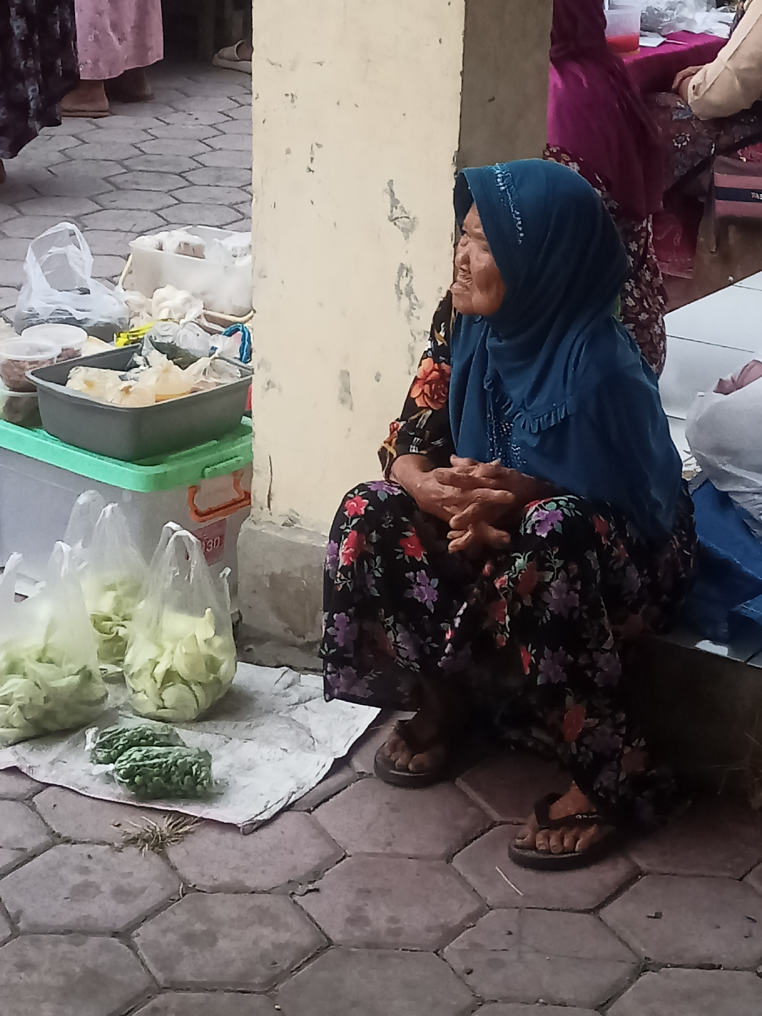 The image depicts an outdoor scene in the city of Nganti, Indonesia, likely a market or roadside stall. An elderly woman wearing a blue headscarf and a floral-patterned dress is seated on a low ledge, facing to her right. Her hands are clasped in her lap. In front of her, on the ground, are several plastic bags filled with what appears to be chopped vegetables, possibly cabbage or lettuce. There are also two smaller packages of green vegetables, possibly chilies or herbs. Beside the woman, to her left, are various containers and plastic bags, suggesting goods being sold or prepared. The ground is paved with hexagonal tiles, and a light-colored pillar is visible to the left of the woman. In the background, other individuals and market stalls are partially visible.