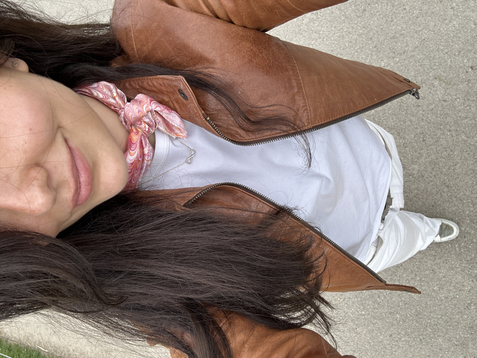 A young woman with long dark brown hair is wearing a brown leather jacket, a white t-shirt, white pants, and a pink and orange bandana. She is wearing a silver infinity necklace and has a faint smile on her face. The woman is standing on a gray paved surface, possibly a sidewalk in the town of Chambord, France, located at latitude: 47.615, longitude: 1.518. She is taking a selfie. The photo appears to be taken from a slightly downward angle. The woman is facing the camera. The background is blurry and contains a few green patches, possibly grass.