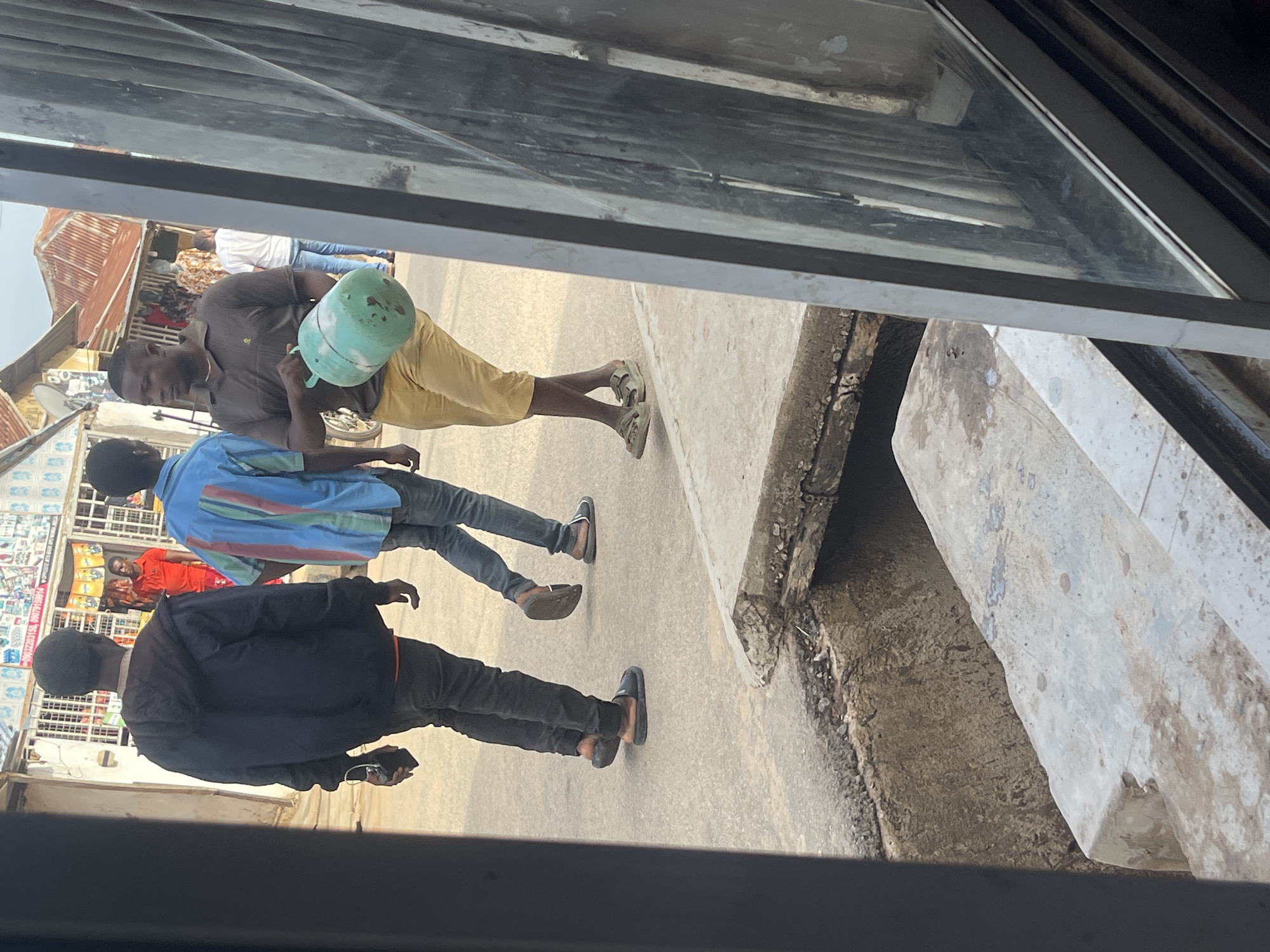 The image depicts an outdoor street scene in Abuja, Nigeria, viewed from behind a dark window frame and glass. The scene features multiple male pedestrians and commercial storefronts under a bright daylight sky.
In the foreground, a dark window frame runs vertically along the left edge, with a reflective glass pane on the right. Below, a concrete curb transitions to a paved surface, revealing an open concrete drain or trench.
Three primary figures are visible in the mid-ground, walking from left to right. The leftmost individual, a male, is seen from behind, wearing a black hooded jacket, dark trousers, and dark slip-on shoes. He carries a dark rectangular object, possibly a mobile phone, in his left hand. To his right, another male is partially visible, also from behind, wearing a vertical striped short-sleeved shirt in blue, red, and green, with blue jeans and dark sandals. The third prominent figure, a male, faces partially towards the viewer, wearing a dark brown polo shirt, tan knee-length shorts, and light-colored sandals. He carries a light green, cylindrical object with a handle, likely a small gas cylinder or water container, in his right hand.
In the background, several single-story commercial buildings line the street. These structures feature visible merchandise displays, including a shop with a red-shirted individual and mannequins dressed in patterned clothing. Some buildings have corrugated metal roofs. The ground surface consists of asphalt road and concrete sidewalks.