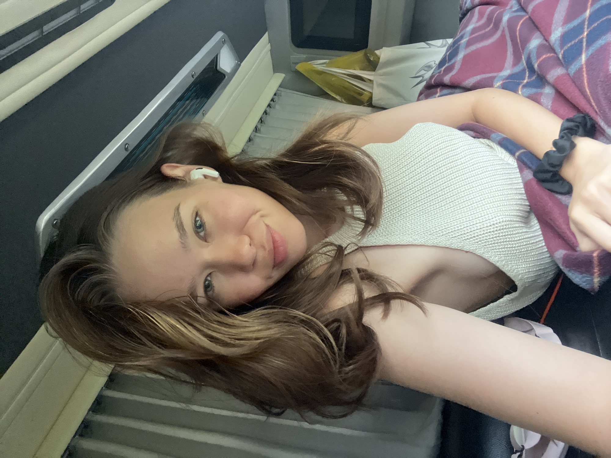 A young woman with light brown hair and blue eyes is shown lying down, taking a selfie. She wears a light-colored, ribbed knit top and has a white wireless earbud in her right ear. A black scrunchie is visible on her left wrist. She is partially covered by a dark red and blue plaid blanket, beneath which a white pillow with a grey design can be seen.
The setting appears to be the interior of a vehicle, possibly a train or bus, suggesting travel. Visible background elements include a silver-framed panel or window, ventilation slats, and storage compartments where a yellow plastic bag and a white bag are stowed. The woman is looking directly at the camera with a calm, soft smile, conveying a sense of relaxation or contentment. The lighting suggests an indoor or enclosed environment, possibly using artificial light. The location context provided for this image is Thôn 1, Vietnam. There is no visible text in the image.