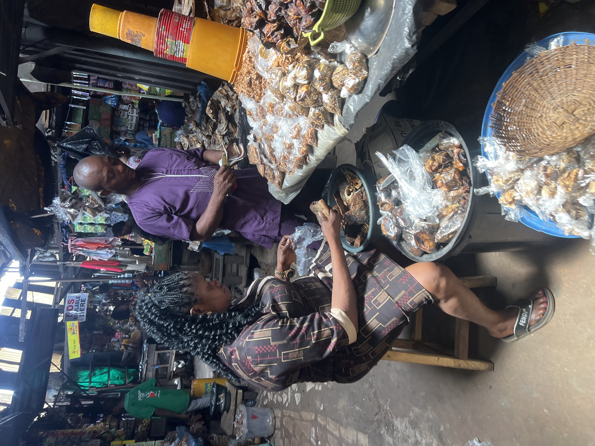 The image shows a market scene in Nkpor, Nigeria. An older man in a purple shirt is standing and counting money. A woman with braided hair wearing a patterned brown dress is sitting on a stool. They appear to be selling dried fish or meat packaged in plastic bags from a stall or vendor station. There is a variety of items displayed in buckets and on tables. The market setting appears somewhat chaotic and busy.