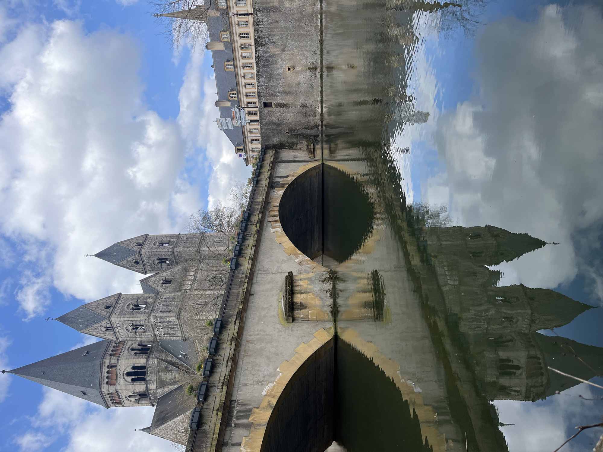 The image shows a reflection of a bridge and a building in a body of water. The building on the left side of the image is the Temple Neuf, which is located in Metz, France. The sky with clouds is reflected on the water's surface. The bridge has an archway visible in the image.