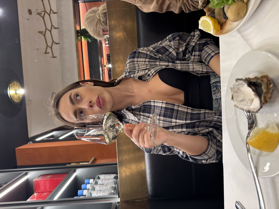A young adult female is seated at a table in an indoor restaurant setting in Verona, Italy. She occupies the left-center of the frame, holding a stemmed wine glass containing a pale yellow liquid in her right hand. Her gaze is directed upwards and to the right, away from the camera, with a neutral expression. She has long brown hair and is wearing a blue and white plaid long-sleeved shirt, unbuttoned, over a black low-cut top. A faint red mark is visible on the upper left side of her chest.
On the white-clothed table in the foreground, a white plate holds a single oyster on the half-shell, a lemon wedge, and a silver fork. Another partially visible plate to the right contains multiple oysters, other shellfish, a lemon wedge, and green garnish.
The background features dark booth seating, a gold-colored partition, and shelves stocked with bottles and red-labeled boxes. A reflective surface behind the woman displays reflections of ceiling lights, an ornate hexagonal light fixture, and the head of another patron with blonde hair tied back. An orange-hued wall section is also visible in the reflection and behind the partition. A partially visible arm in a brown garment belonging to a second individual is seen to the right of the main subject.