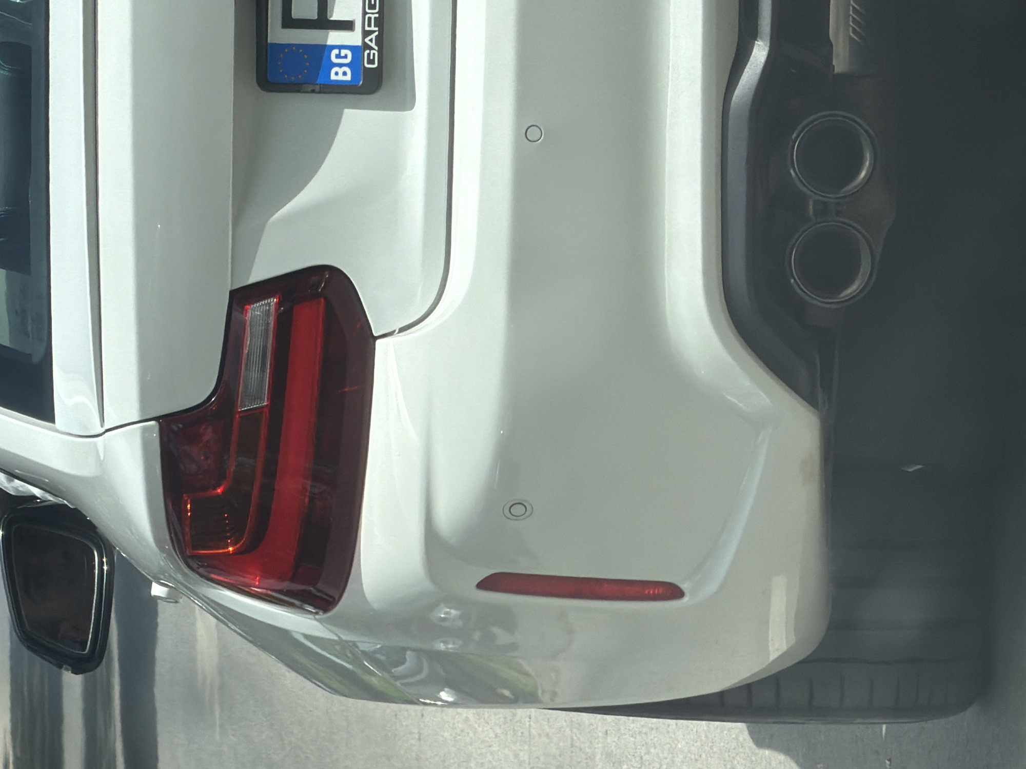 The image shows the rear of a white car with a Bulgarian license plate ("BG"). The car features a modern taillight design, parking sensors, and dual exhaust pipes. The location is suggested to be in Plovdiv, Bulgaria. The vehicle appears to be parked.
