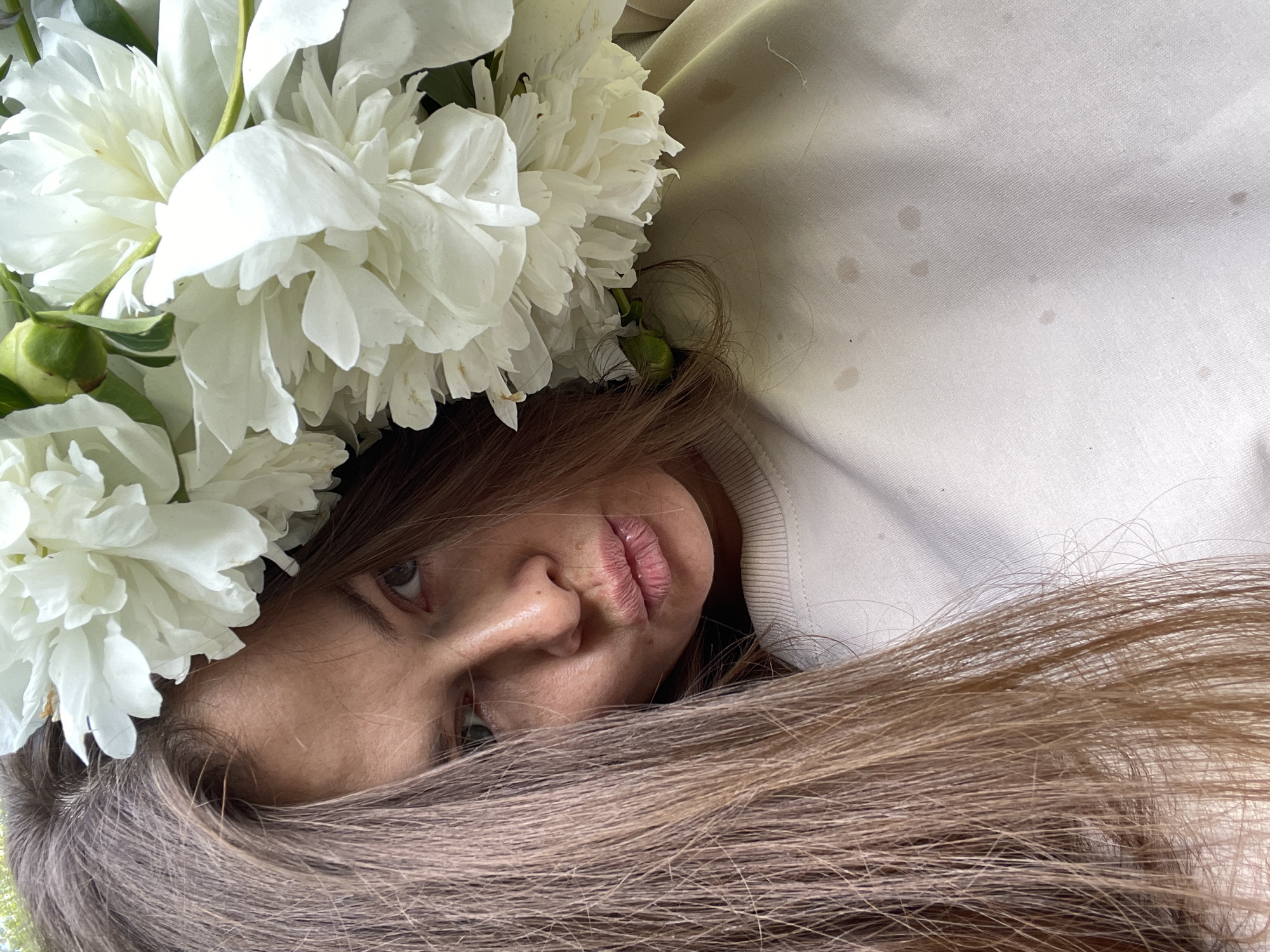 The media file shows a close-up, angled shot of a young woman lying down, wearing a crown of white peonies. The woman's face is partially obscured by her long, light brown hair and the flowers. Her eyes are visible, looking towards the camera, and her lips are slightly parted. She appears to be indoors, or in a shaded outdoor area, as indicated by the soft lighting and the presence of what looks like a light-colored fabric garment with subtle stains. The background is a plain, light-colored fabric, possibly a blanket or clothing. The overall mood of the image is serene and perhaps a little whimsical due to the floral crown. There is no visible text in the image. The location context is Заречный, Russia.